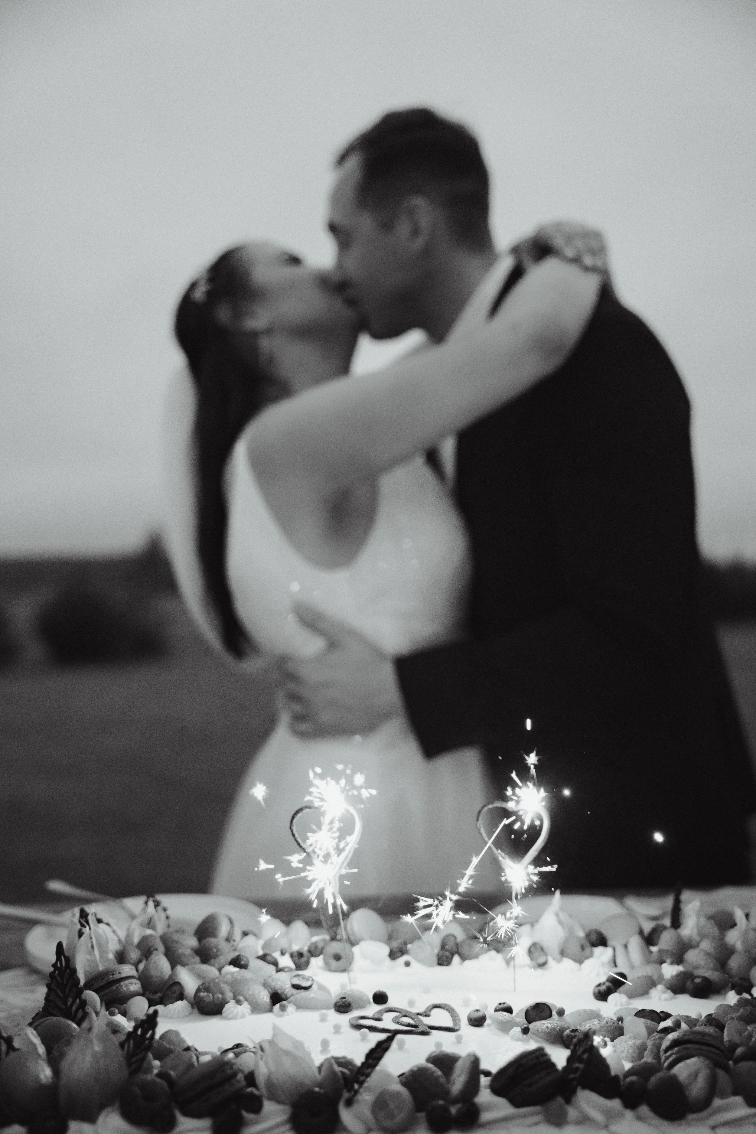 A black-and-white photo of a couple kissing in front of a large cake decorated with candies and sparklers.