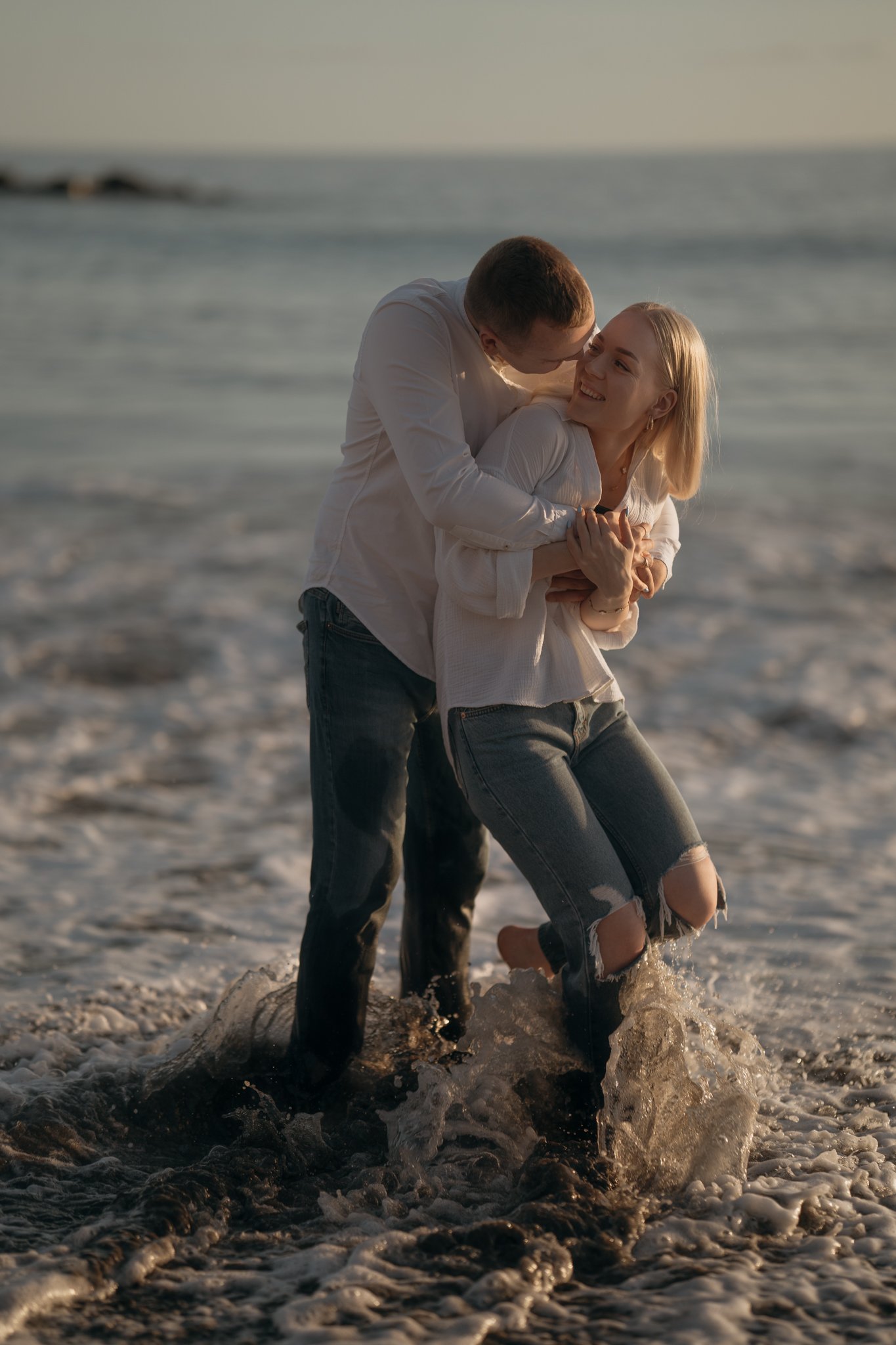 A couple at the beach, with the man lifting the woman as they play in the shallow water near the shoreline during sunset.
