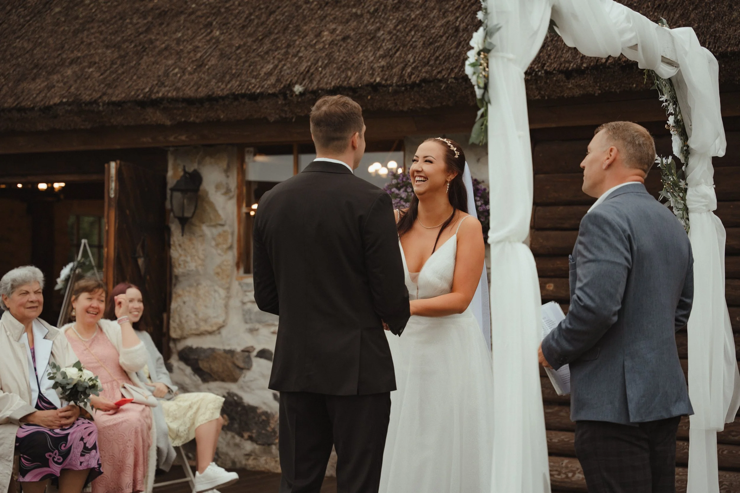 Bride and groom exchanging vows during wedding ceremony under white decorated archway, with guests watching and smiling in the background.
