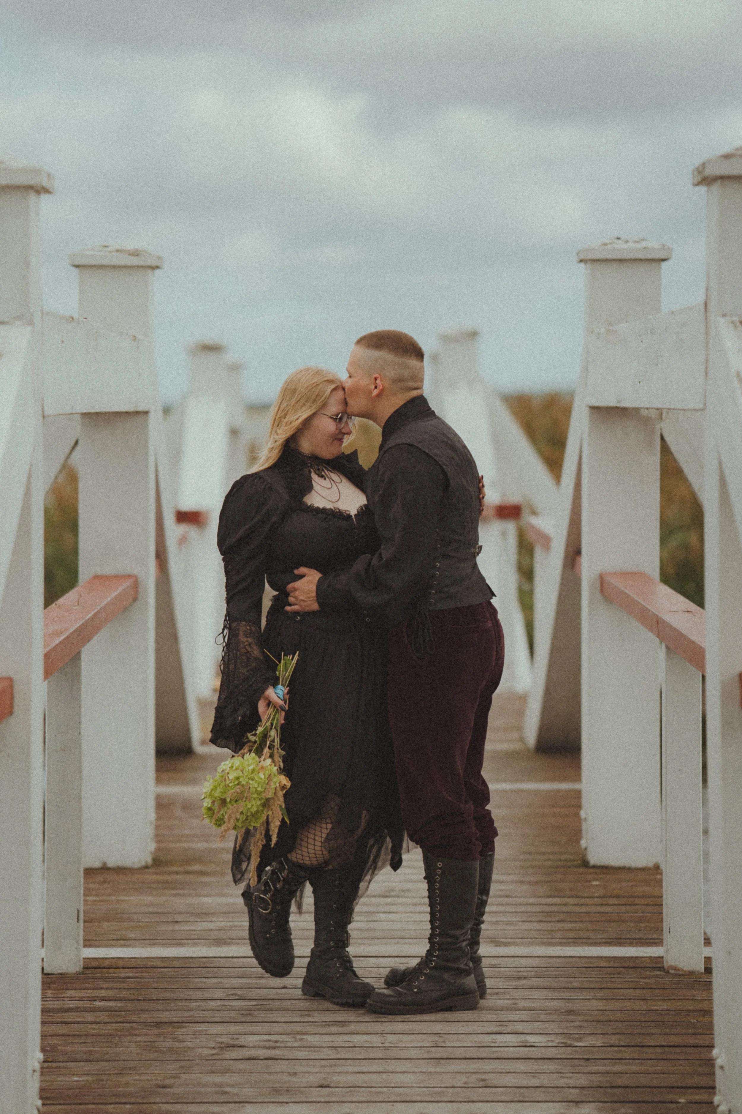 A couple dressed in goth attire sharing a kiss on a wooden bridge with a cloudy sky in the background.