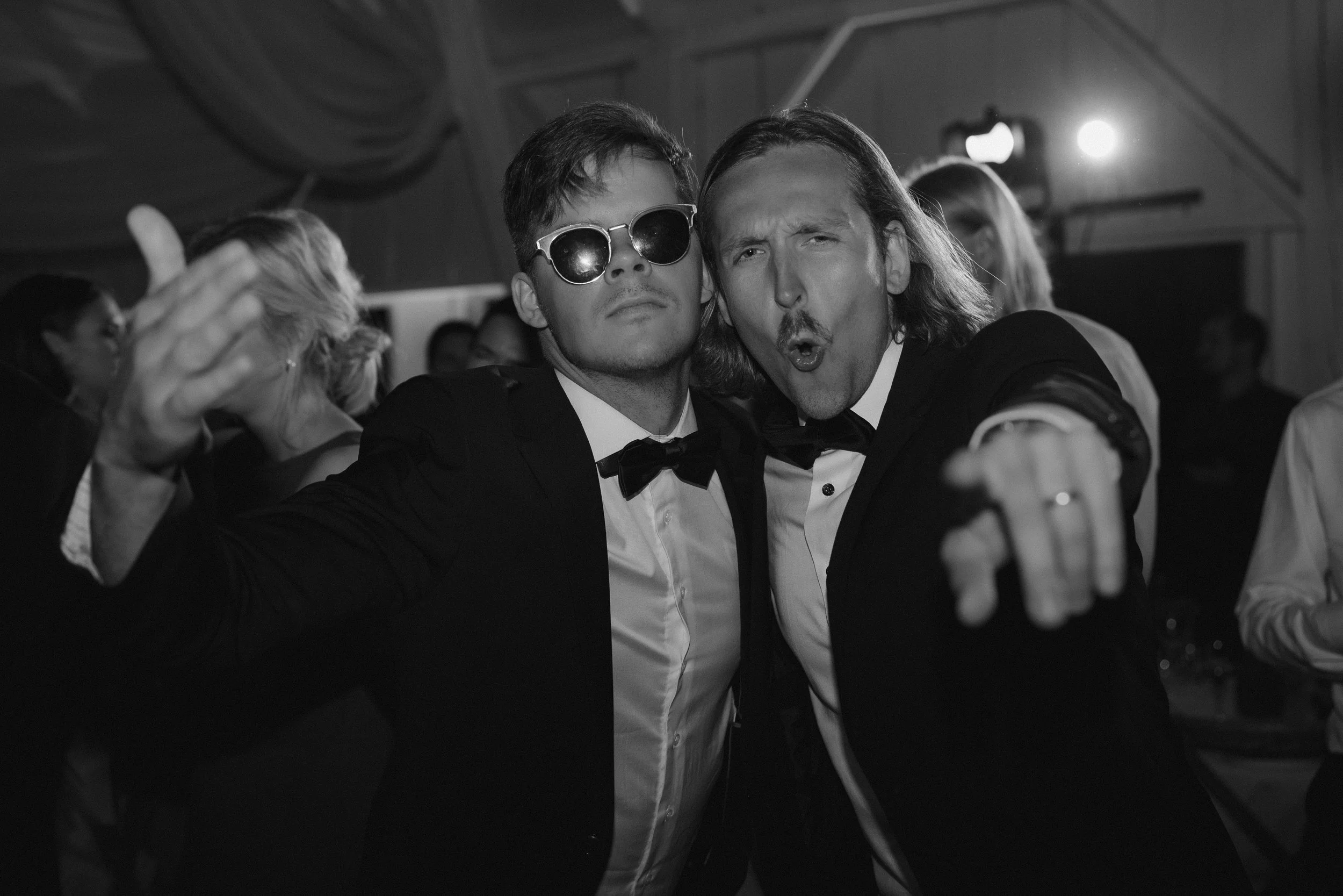 Two men in tuxedos at a party, one with sunglasses, making expressive faces, with other people in the background.