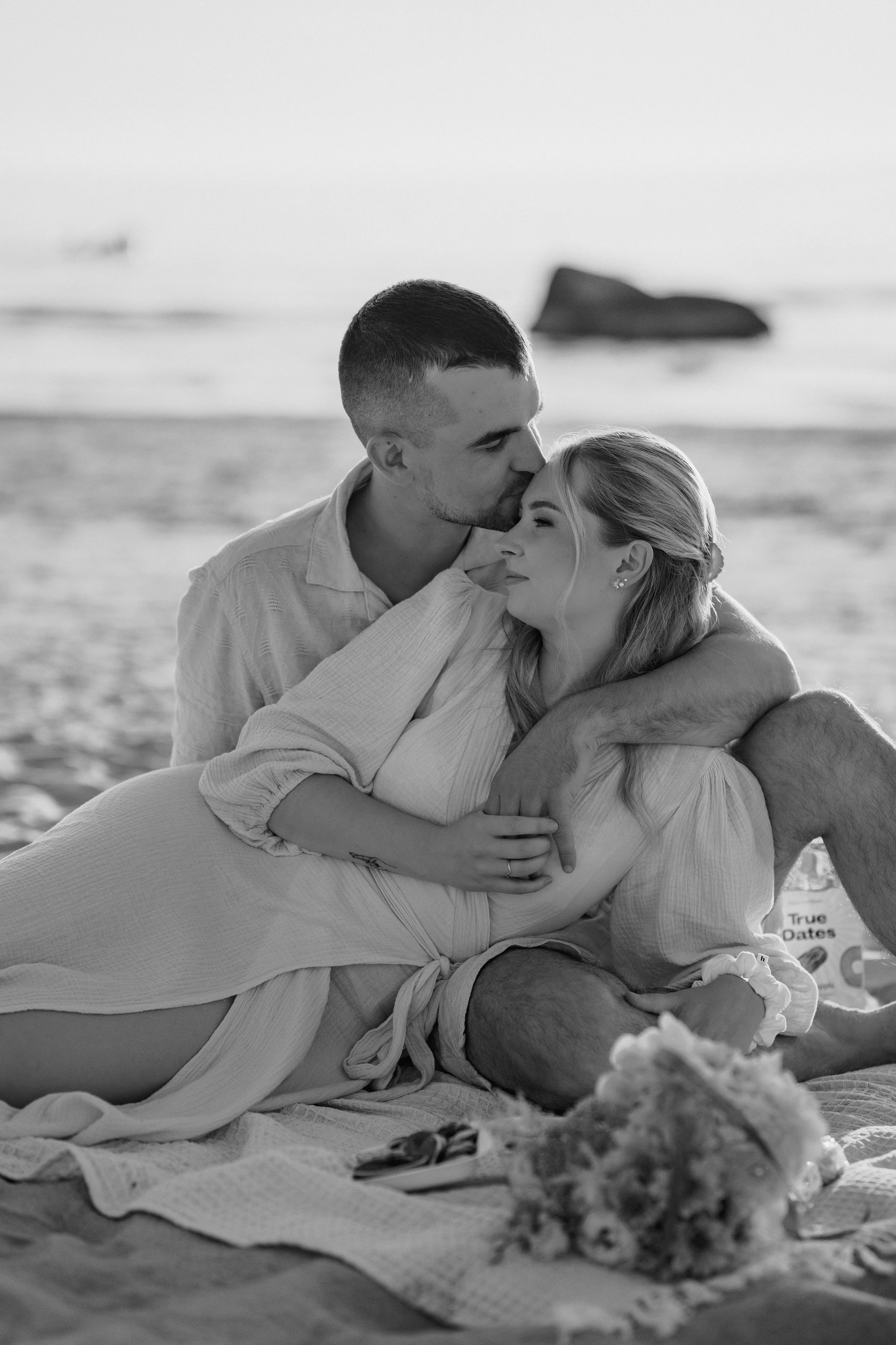 A Black and white photo of a couple sitting on a blanket at the beach, with the man kissing the woman's forehead. The woman is smiling and resting her hands on the man's arm. They are surrounded by a peaceful beach setting with the ocean and rocks in the background.