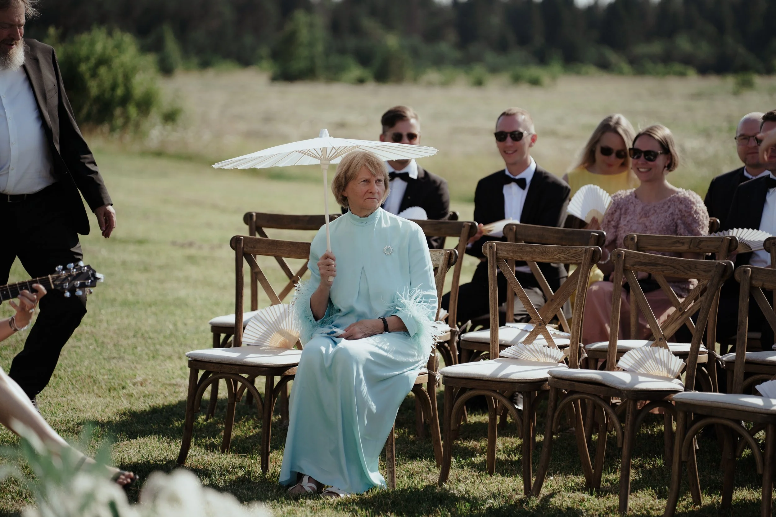 Older woman in a mint green dress sitting on a wooden chair outdoors, holding a white parasol, at a formal event with other seated guests in tuxedos and dresses, some holding fans, in a grassy area with trees in the background.