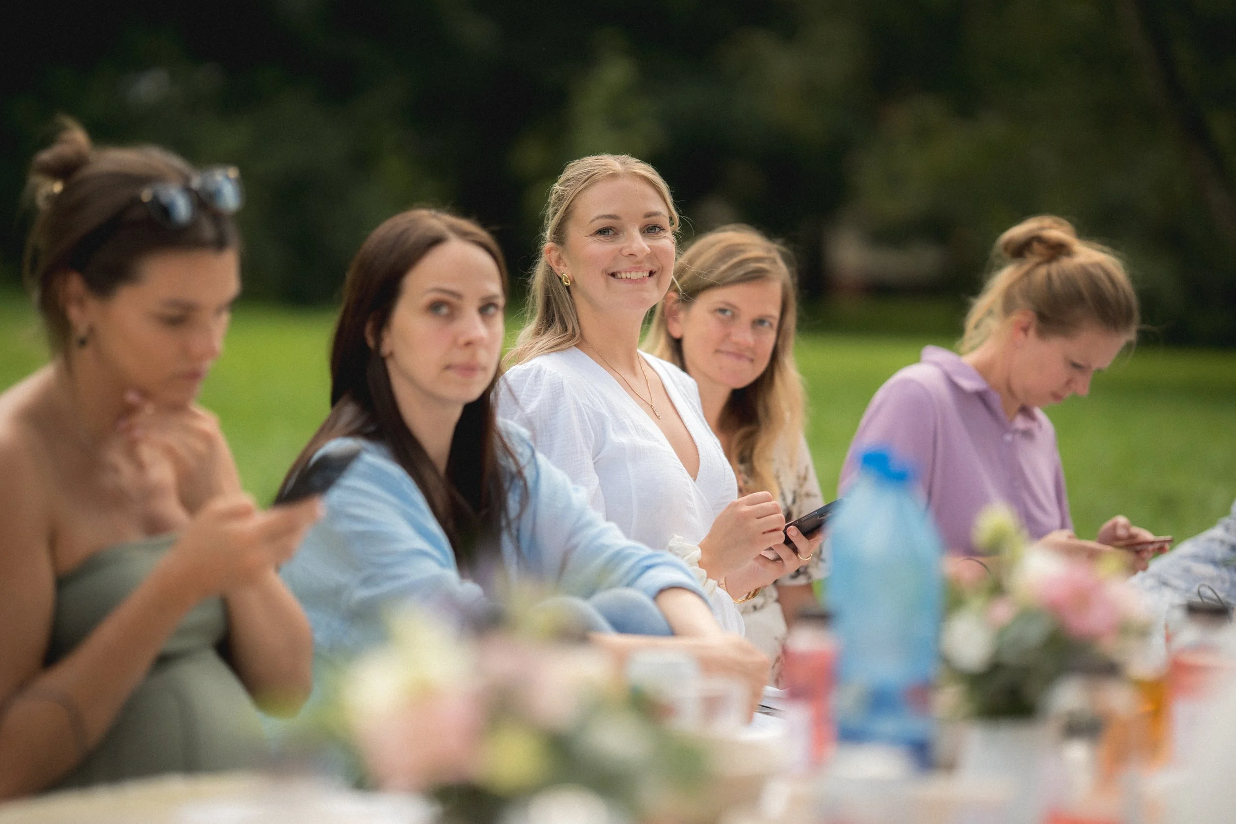 A group of women sitting outdoors at a table during daytime, some using smartphones, with a woman smiling at the camera in the center. There are blurred decorations and water bottles on the table.