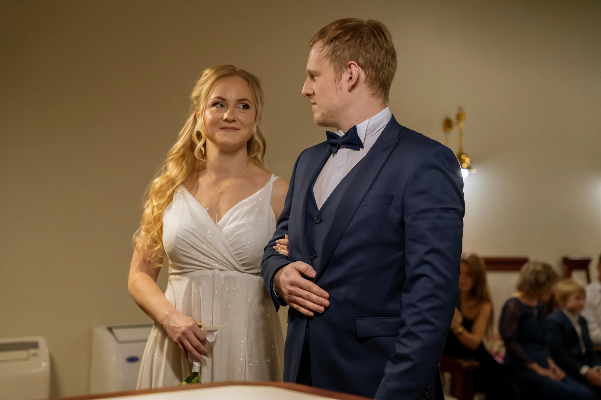 A woman in a white dress and a man in a blue tuxedo are standing close to each other indoors during a formal event. The woman is looking at the man with a smile, and the man is looking at her with a serious expression.