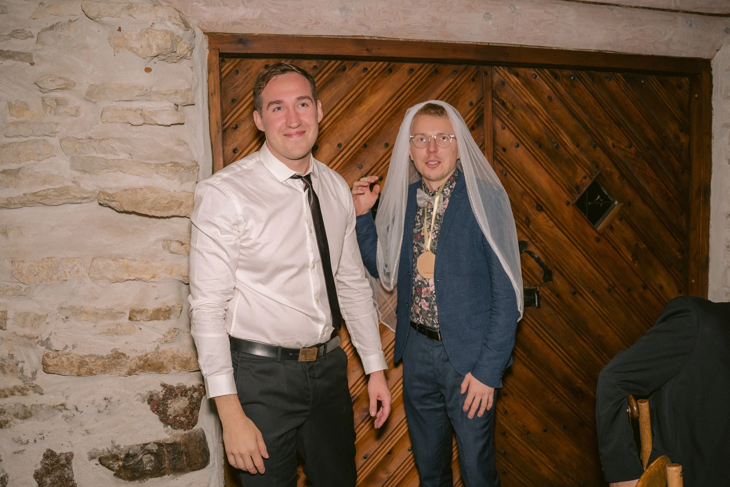 Two men standing indoors, one wearing a white shirt and black tie, the other wearing a blue suit with a floral shirt, glasses, and a veil, suggesting a humorous or wedding-related context, against a stone and wooden wall background.