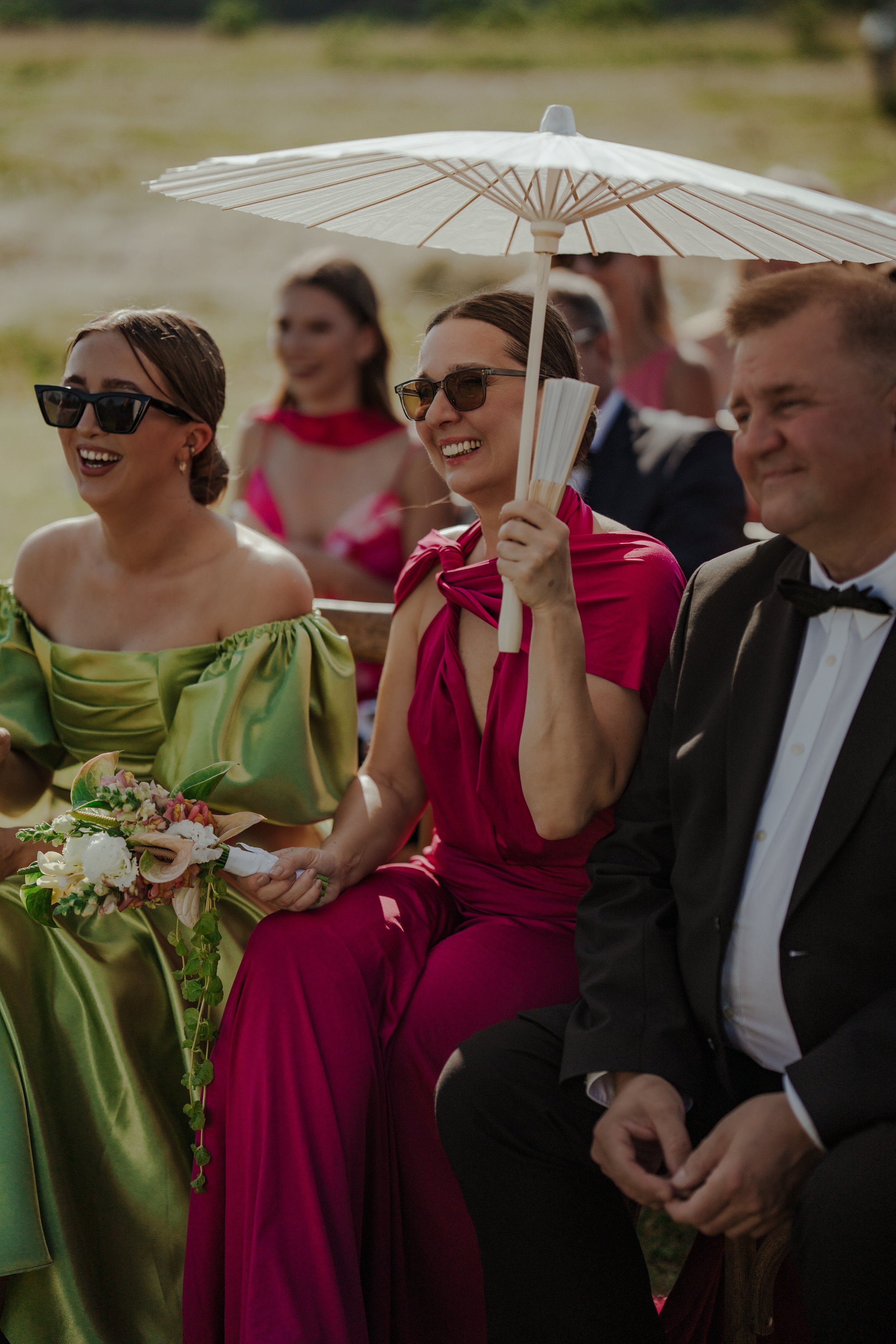 Group of people dressed in formal attire, sitting outdoors, smiling and enjoying a sunny day, with one woman holding a parasol and a bouquet of flowers.