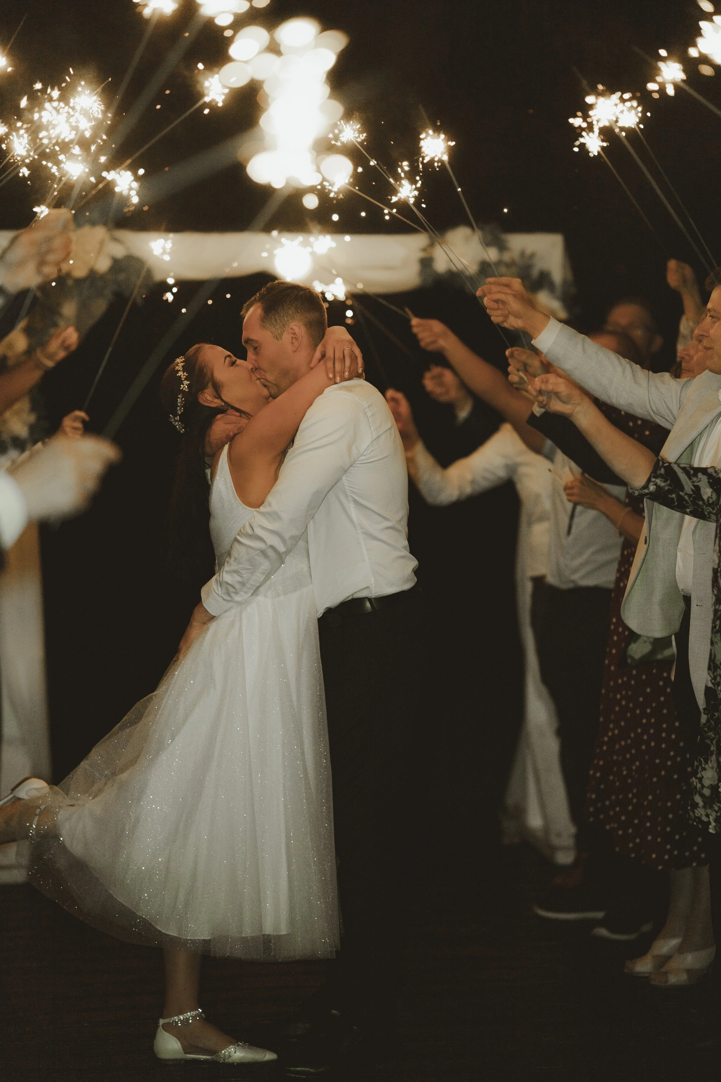 A bride and groom kiss while their wedding guests celebrate with sparklers during their wedding reception at night.