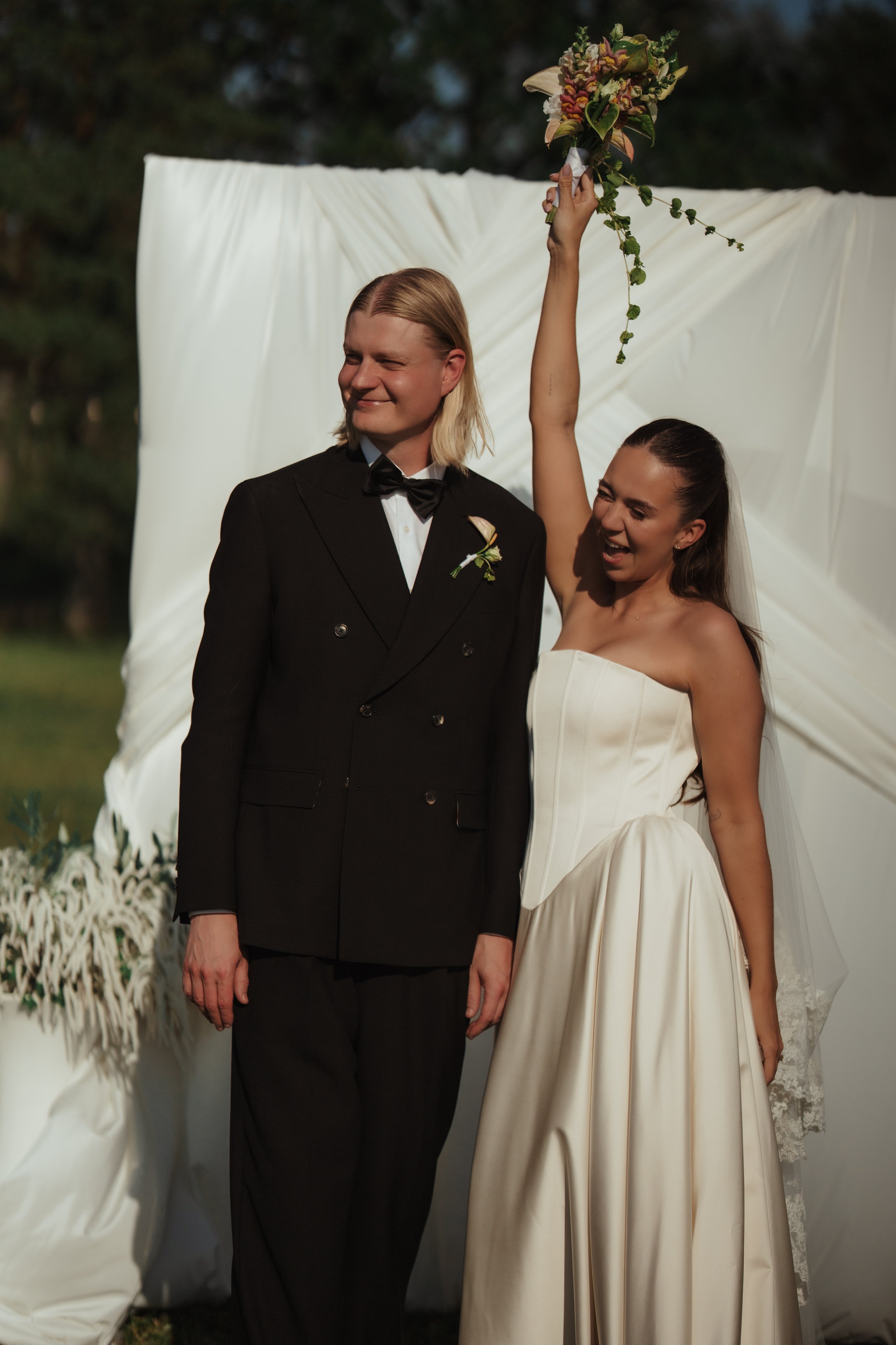 A bride and groom celebrating their wedding outdoors, standing in front of a white fabric backdrop. The bride, in a strapless white wedding gown, holds a bouquet high in the air and laughs, while the groom, in a black tuxedo, stands beside her smilin