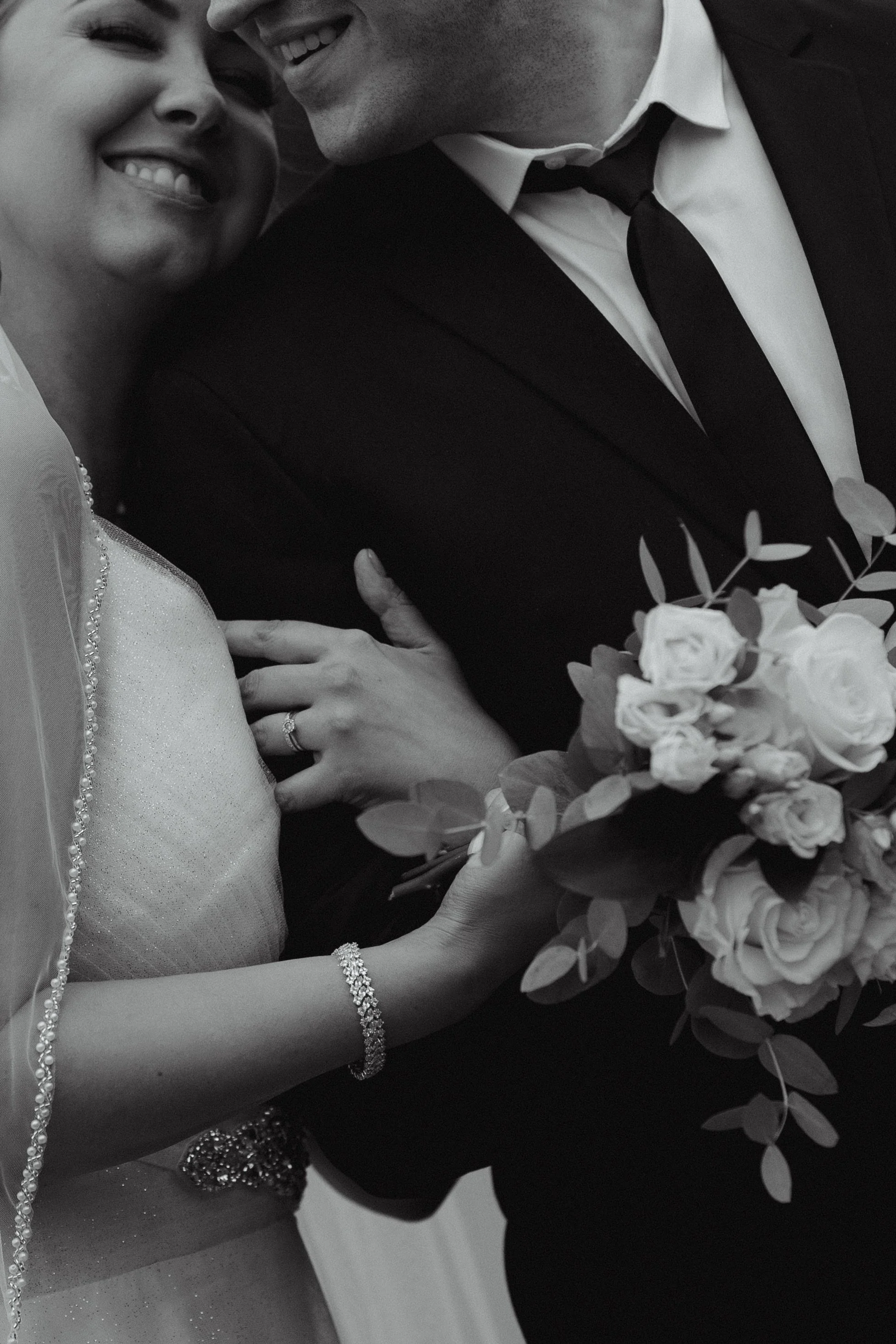 Black and white photo of a smiling bride and groom close together, with the bride holding a bouquet of roses and greenery, and showing her jewelry.