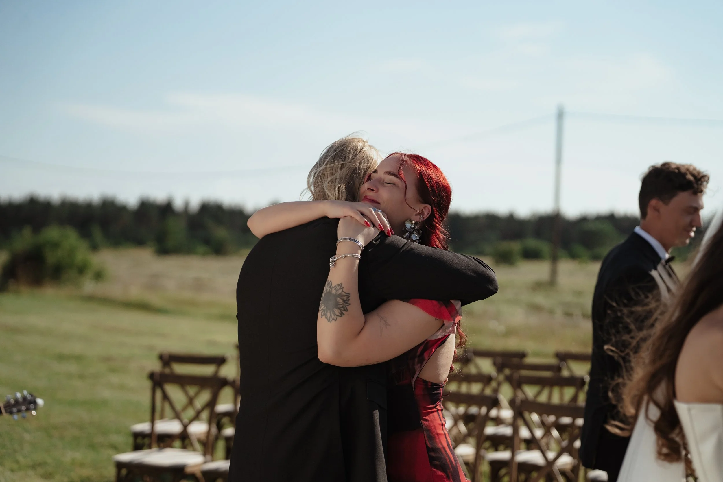 Two women embracing warmly outdoors at a celebration or event, with chairs and other people in the background.