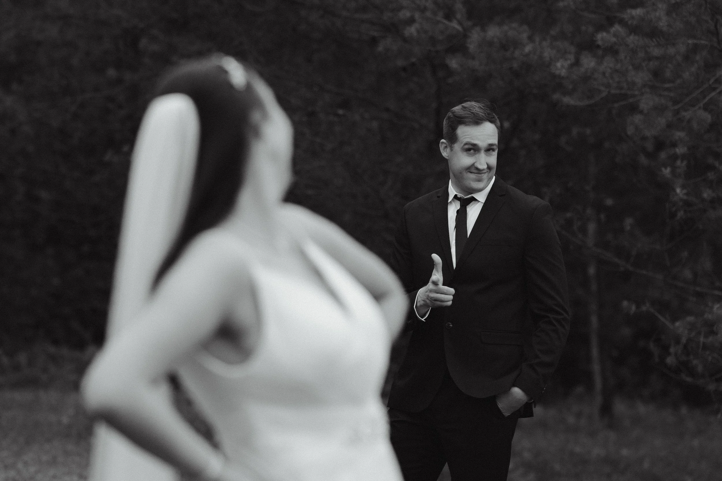 A black and white photo of a man in a suit giving a thumbs-up and smiling, while a woman in a white dress stands in the foreground with her back to the camera, outdoors near trees.