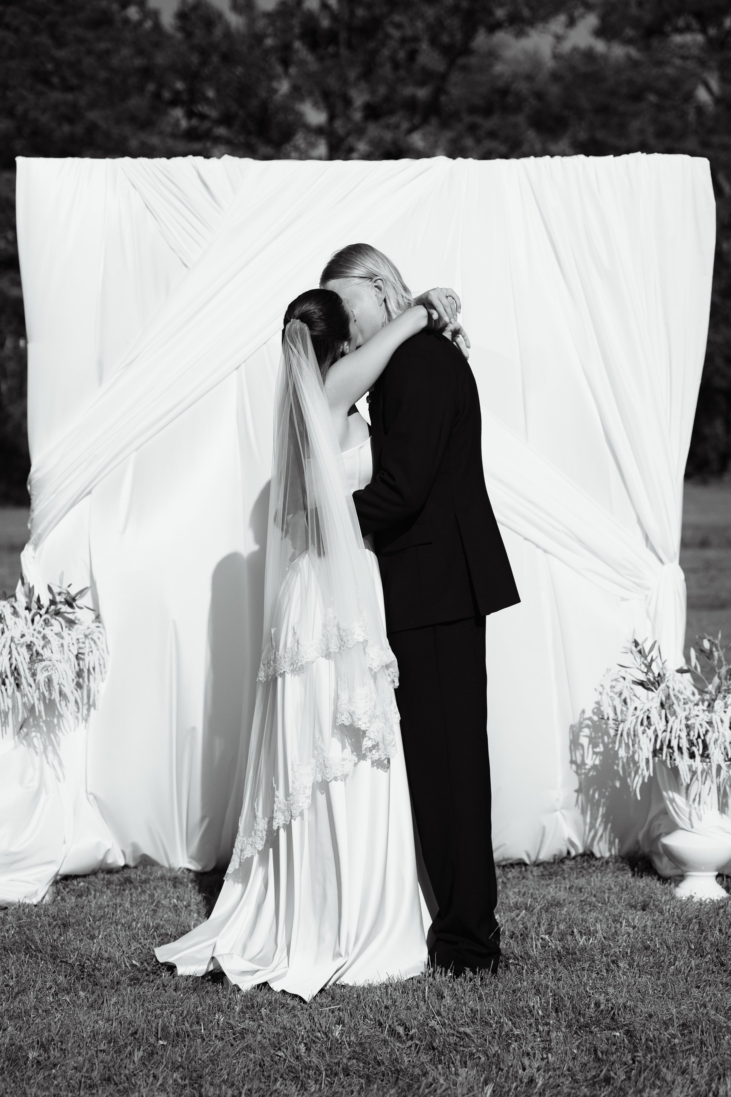 A black and white photo of a bride and groom sharing a kiss at their wedding ceremony outdoors. The bride is wearing a white dress and veil, and the groom is dressed in a black suit. They are standing in front of a draped white backdrop with floral a