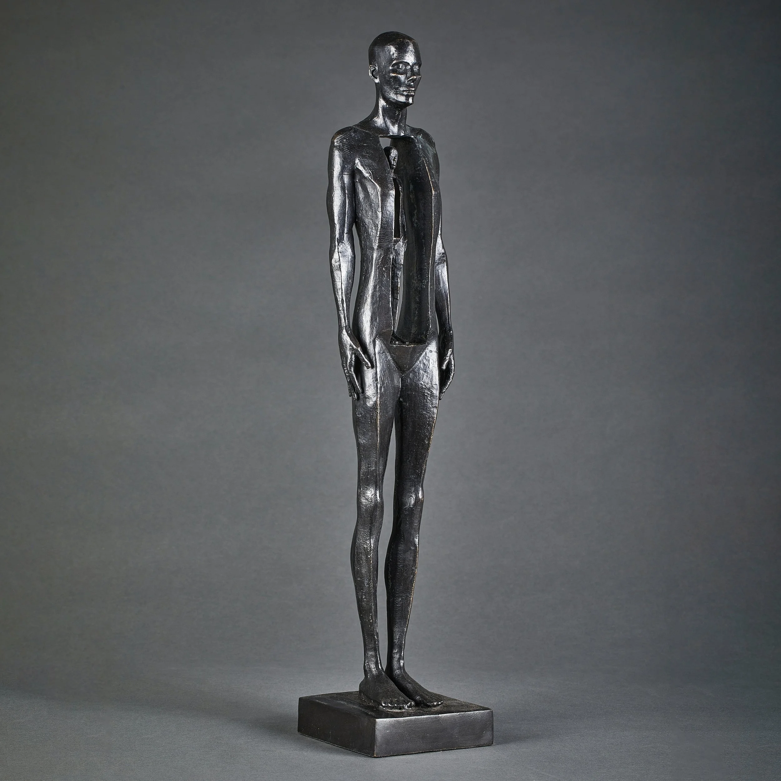 A black metallic sculpture of a tall, slender human figure with an elongated body and stylized features, standing on a rectangular base against a plain grey background.