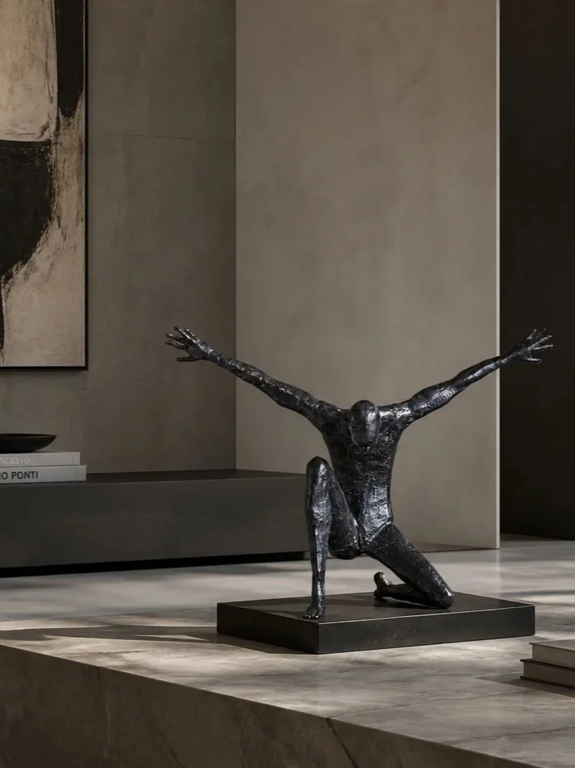 A black sculpture of a person in a kneeling lunge position with arms extended outward and upward, placed on a black rectangular base in a modern interior setting.