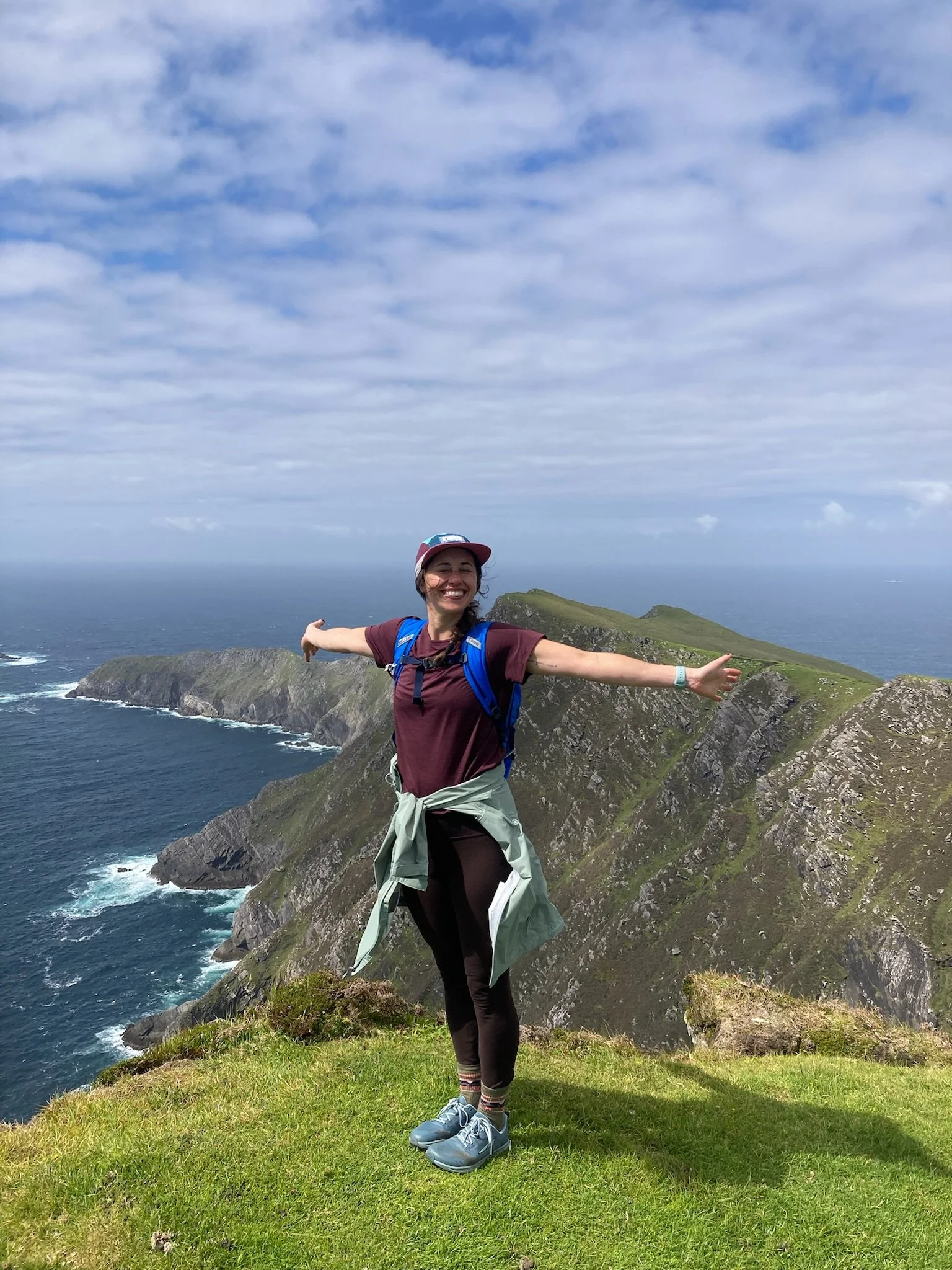 A woman smiling and spreading her arms wide on a grassy cliff overlooking the ocean, with cloudy sky above and rugged cliffs in the background.