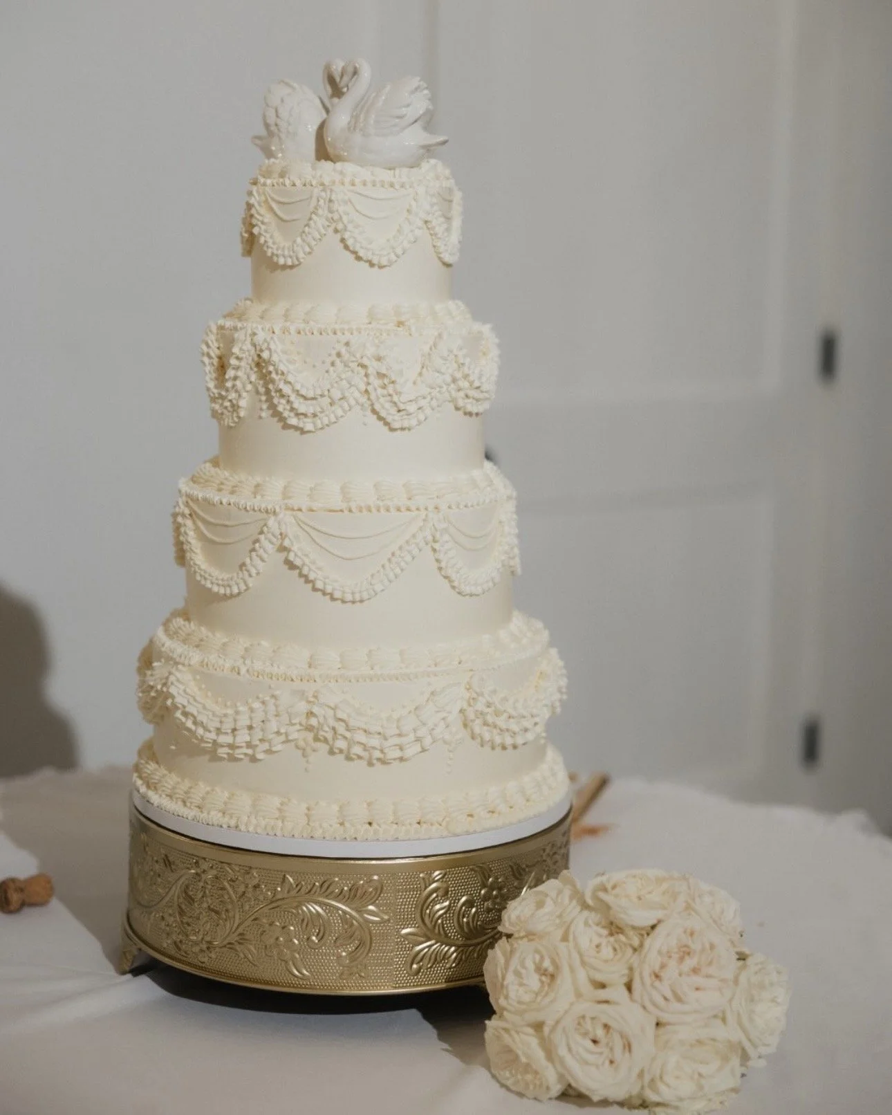 A tall four-tiered white wedding cake with intricate piping and decoration, topped with a swan figurine. The cake sits on a decorative gold base, with a bouquet of white roses nearby.
