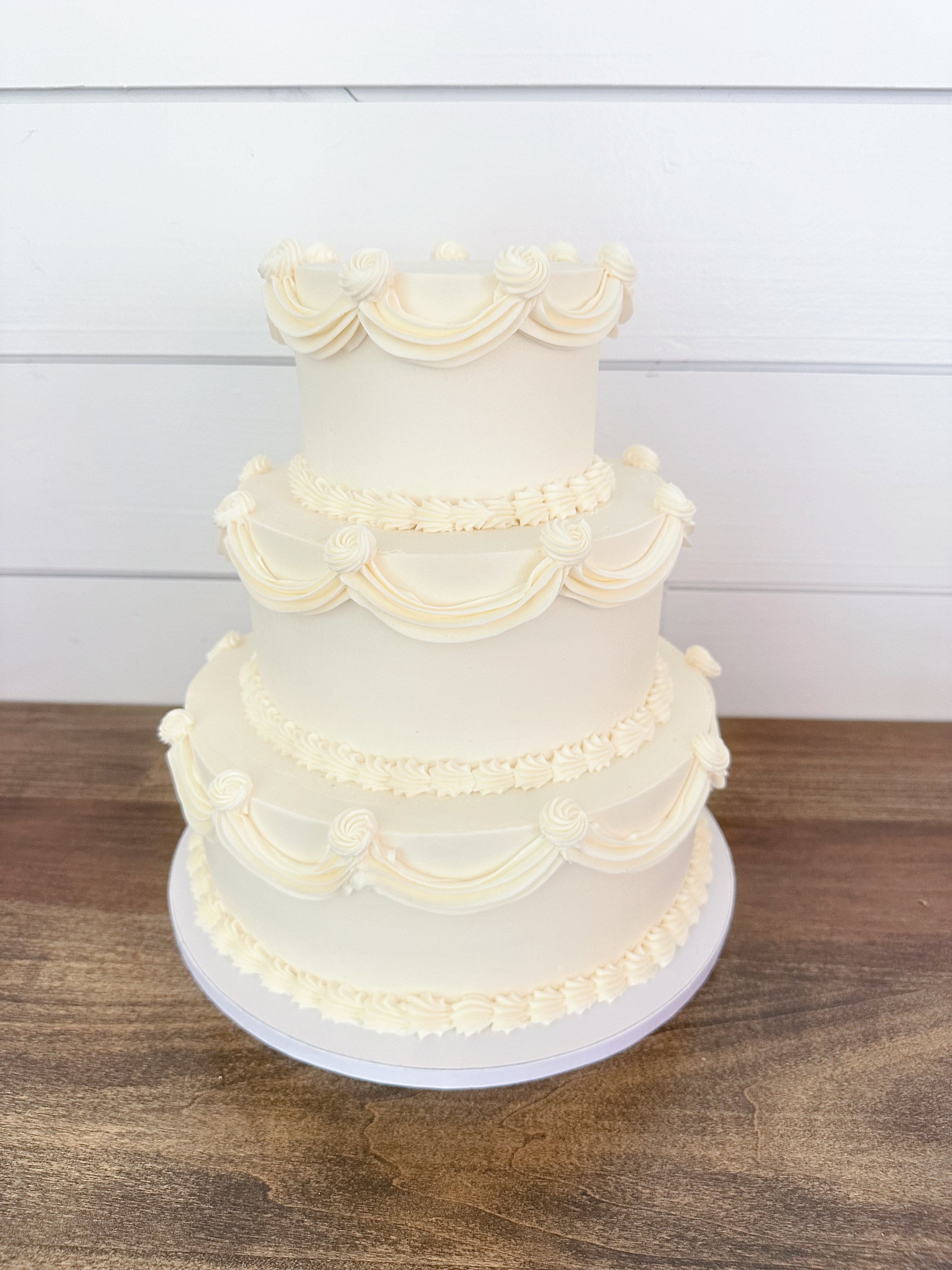 Four-tiered white wedding cake with piped cream decorations, placed on a white cake board on a wooden surface, against a white paneled background.