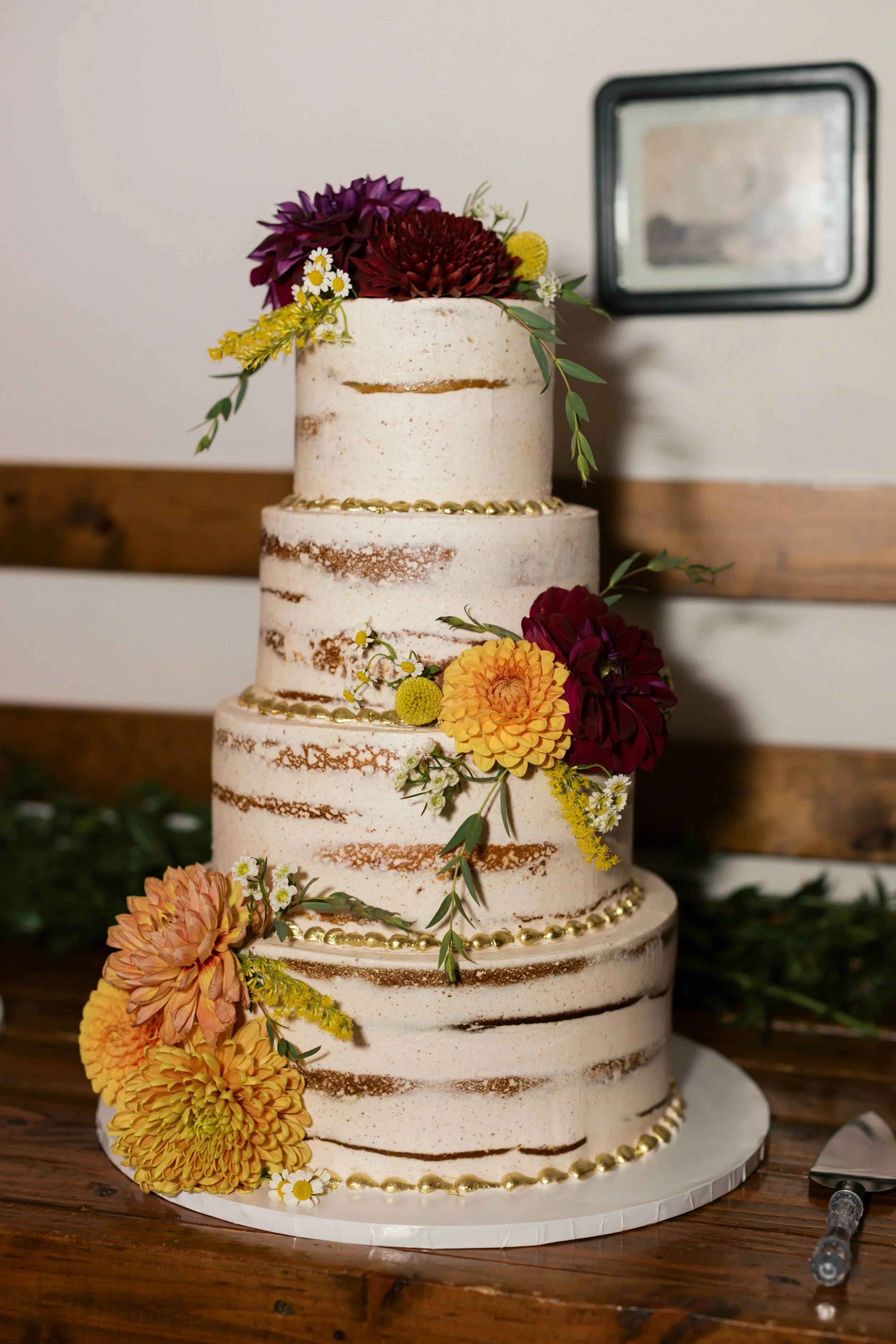 A multi-tiered wedding cake with semi-naked frosting, gold decorative beads, and vibrant flowers