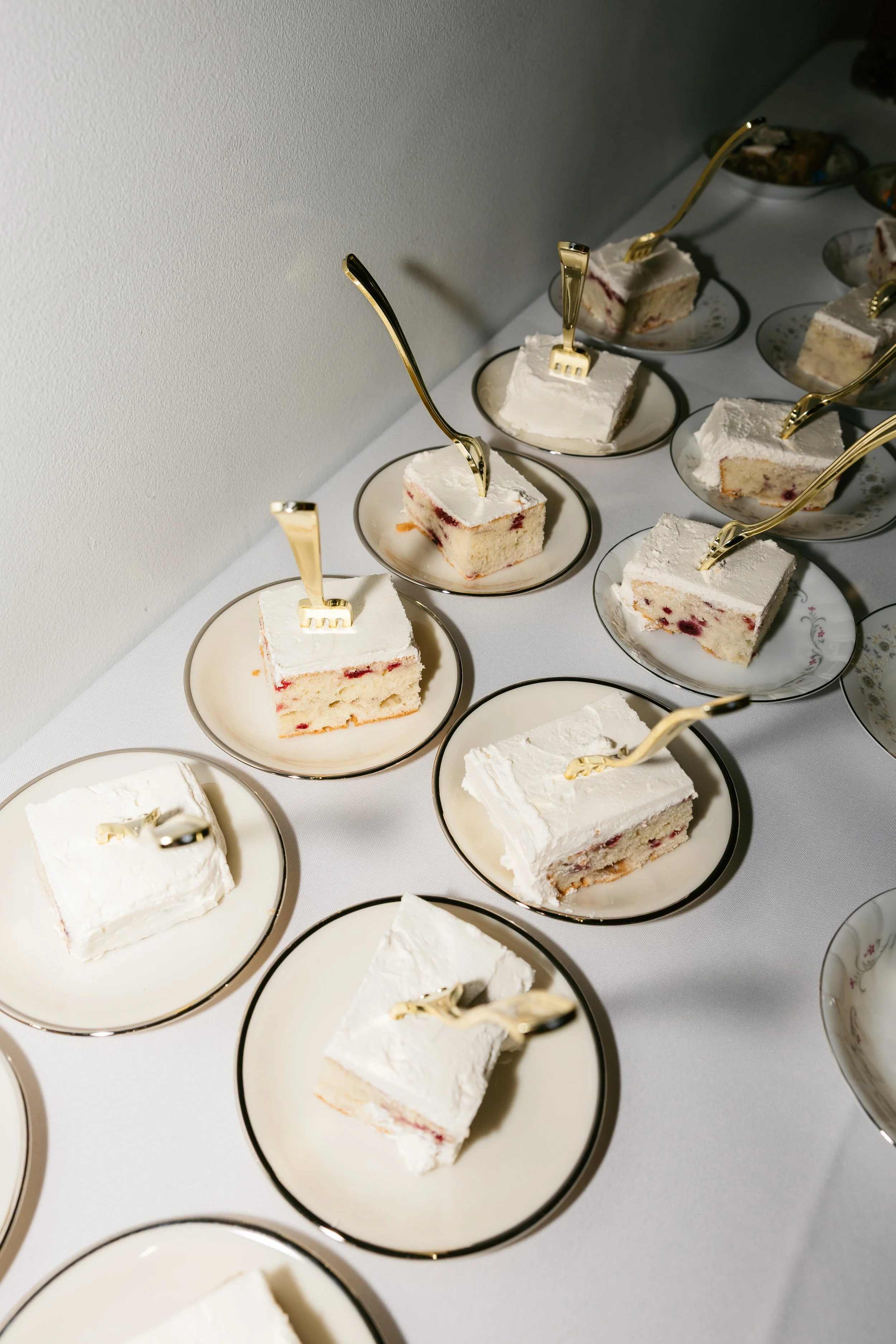 Assorted slices of cake with white frosting and a gold fork on each plate, arranged on a white tablecloth.