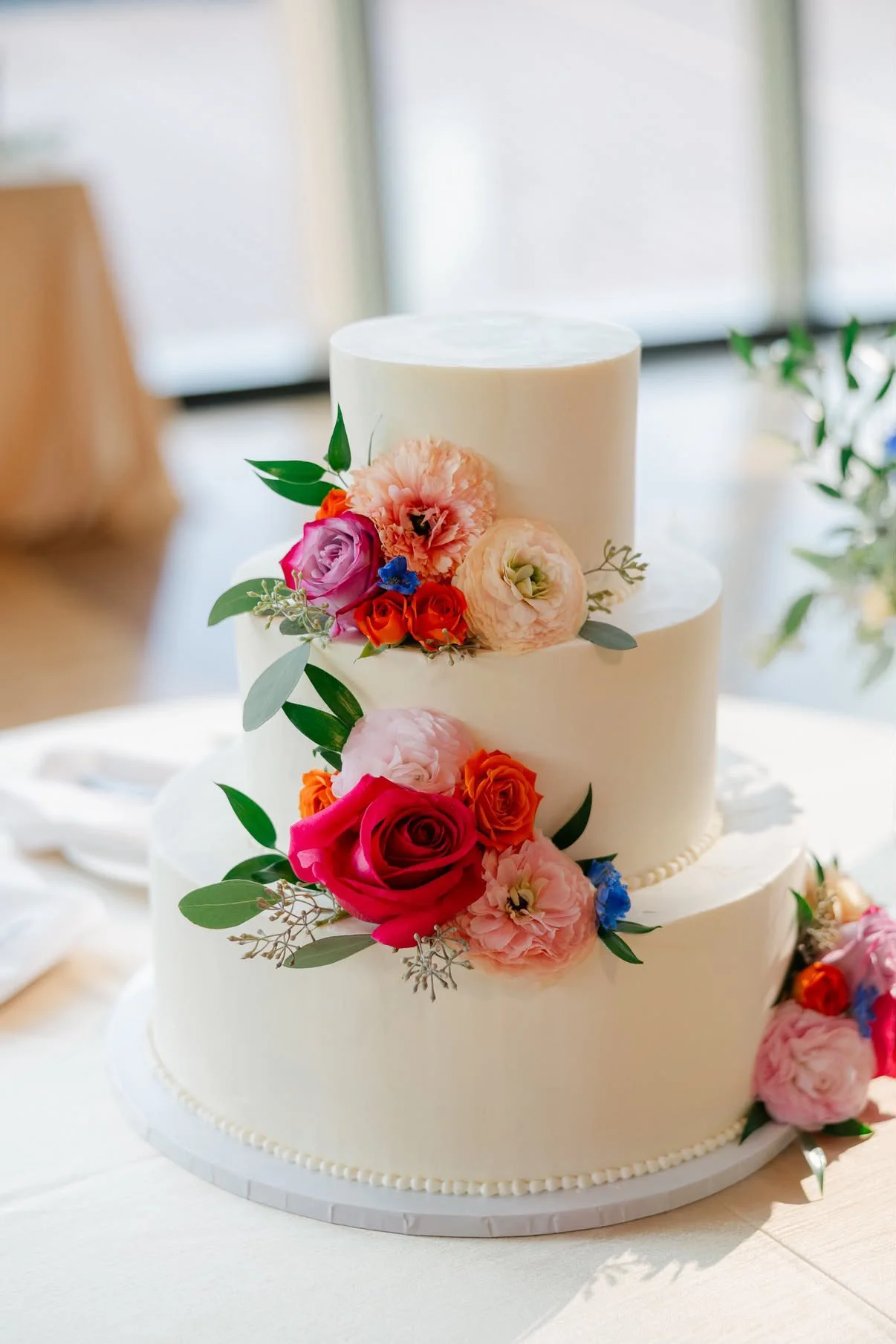 A three-tier white wedding cake decorated with pink, purple, orange, red, and blue flowers and green leaves.