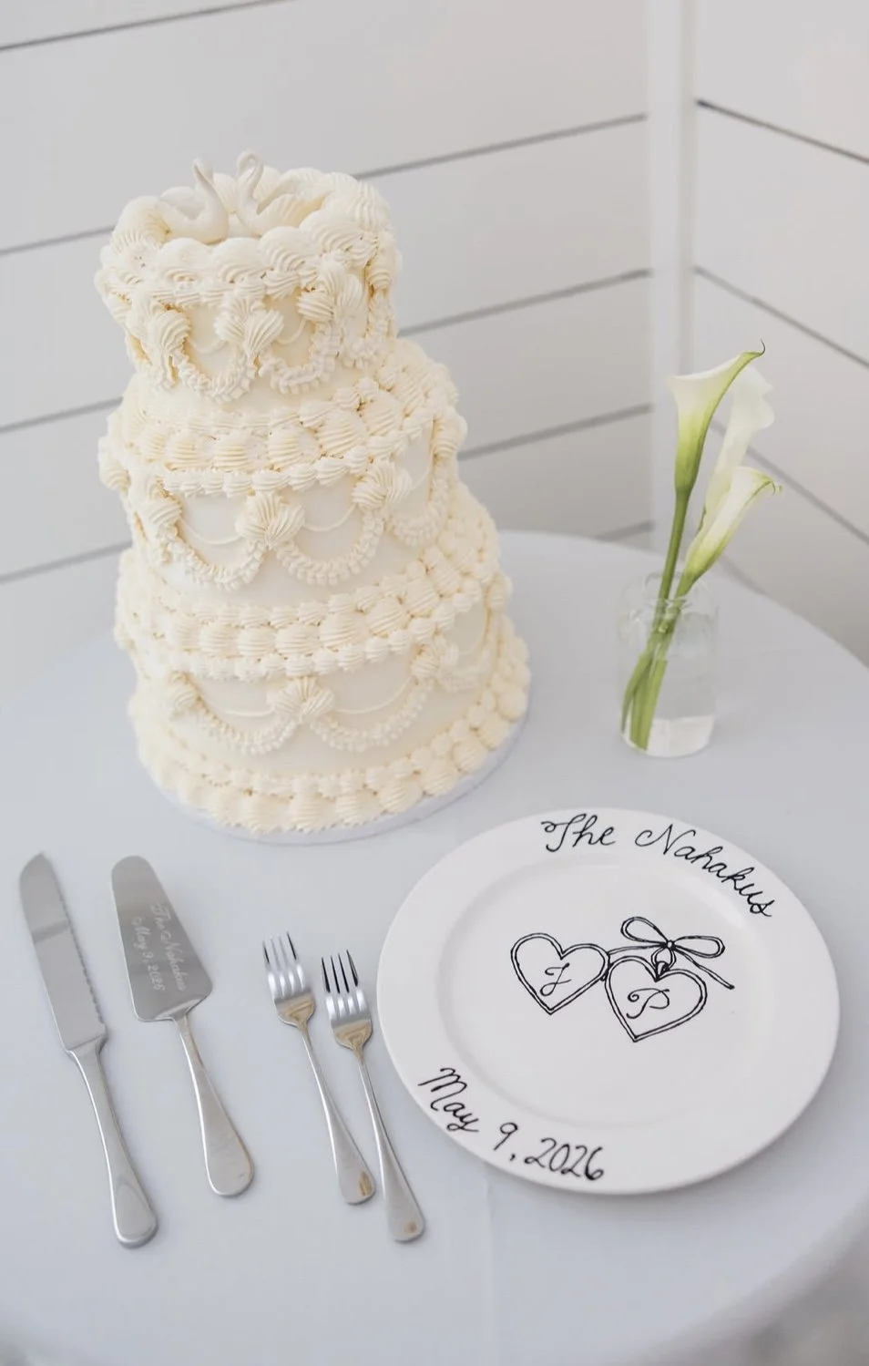 A wedding cake with white icing and intricate piping decorations, a white plate with a heart and bow design, a clear vase with white calla lilies, and silver utensils on a white tablecloth.
