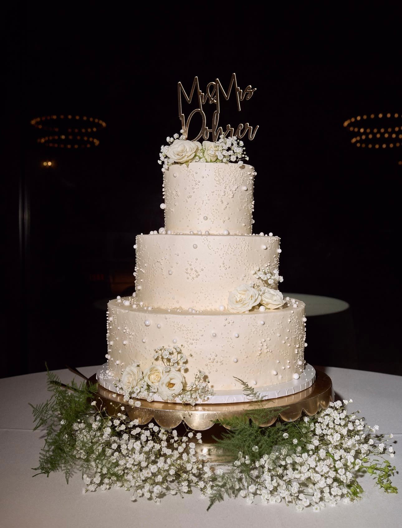 Elegant three-tier white wedding cake decorated with white flowers and small white pearls, topped with a gold 'Mr & Mrs Parker' cake topper, on a silver stand, surrounded by greenery and baby's breath flowers.