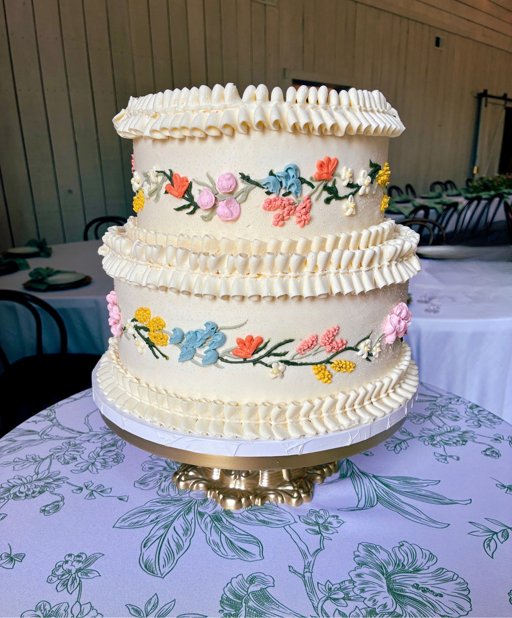 Multi-tiered white wedding cake decorated with colorful floral piping, placed on a decorative cake stand on a table with a floral tablecloth.