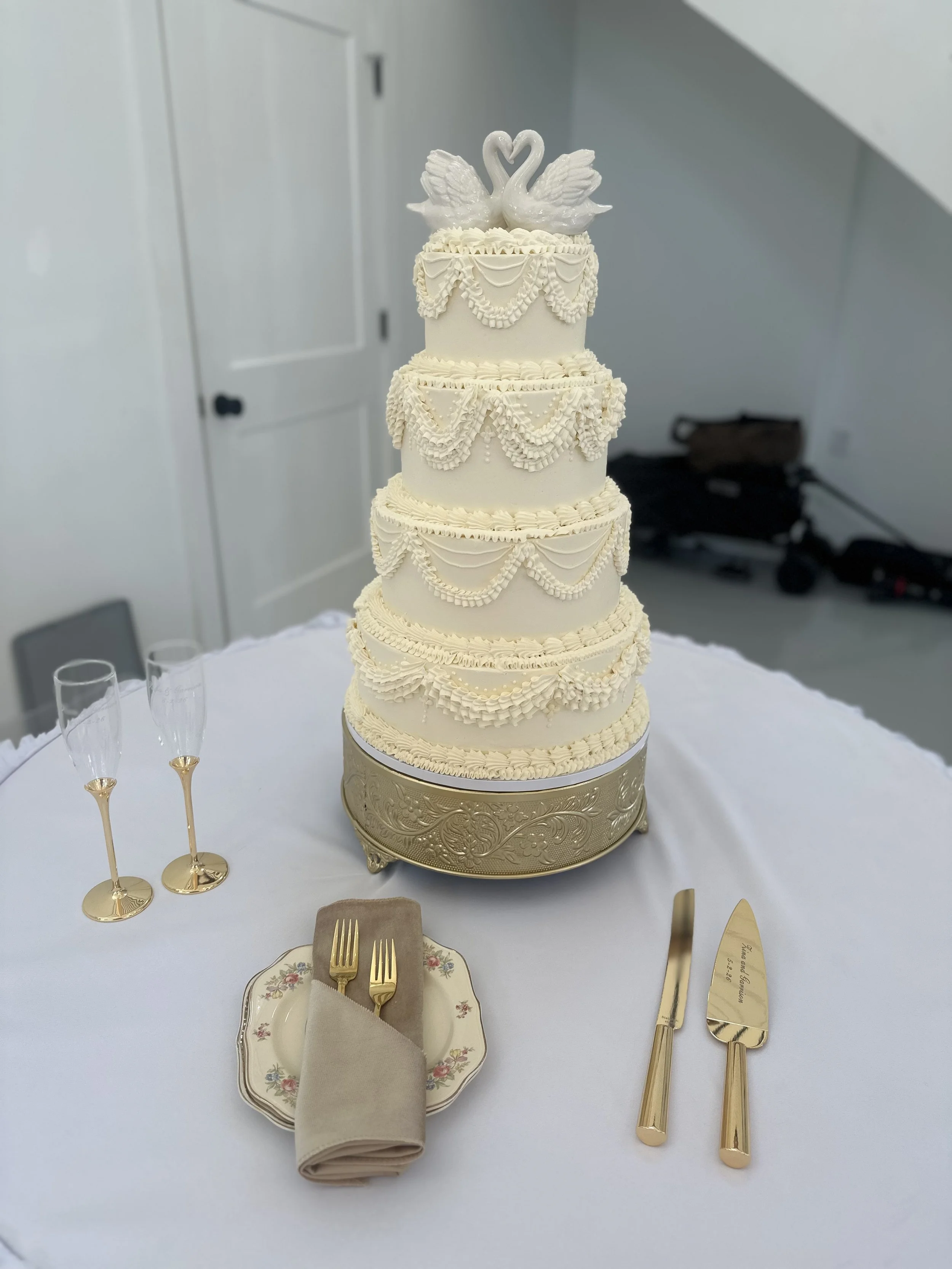A tall, tiered wedding cake decorated with white icing, intricate piped designs, and two swan figurines on top, sitting on a gold ornate cake stand. The cake is surrounded by champagne flutes and tableware on a white tablecloth.