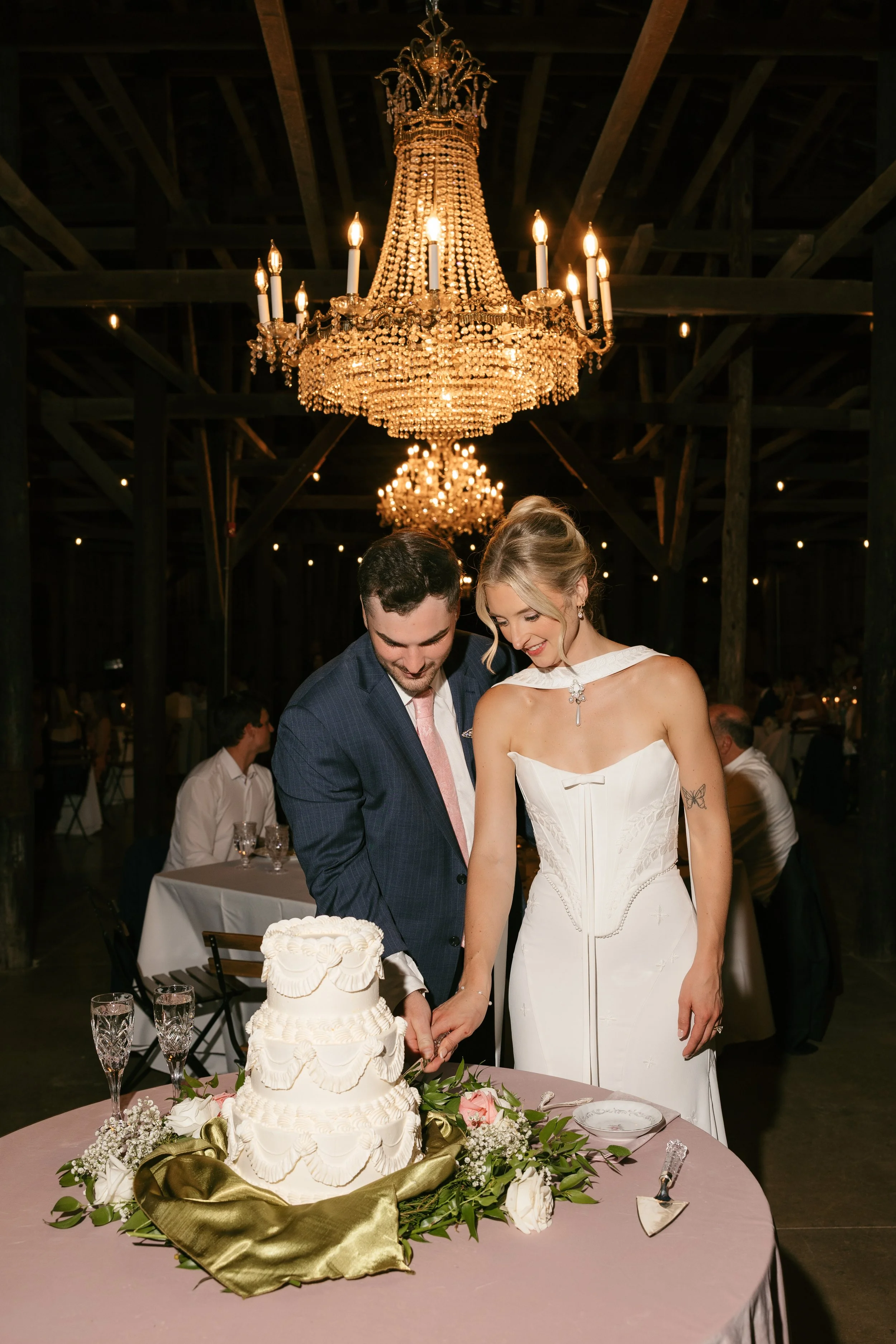 A bride and groom cutting a wedding cake at their reception, with a crystal chandelier hanging above them and guests in the background.