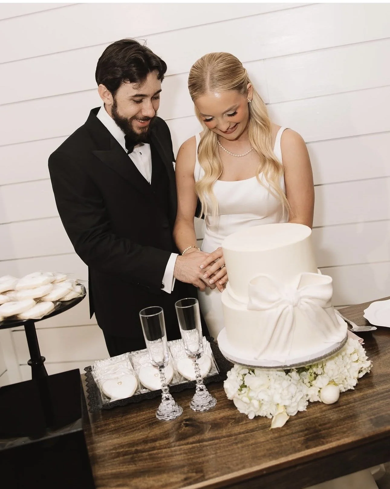 A bride and groom cutting a white wedding cake decorated with a bow, surrounded by wedding desserts and champagne glasses at a celebration