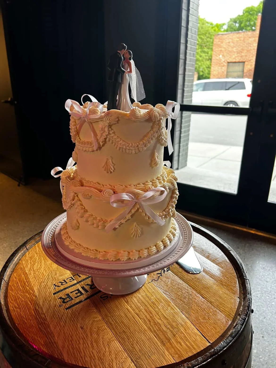 A two-tier wedding cake decorated with pink ribbons and piped white icing, topped with a bride and groom figurine, placed on a round wooden table near a door with an outdoor scene visible through the glass.
