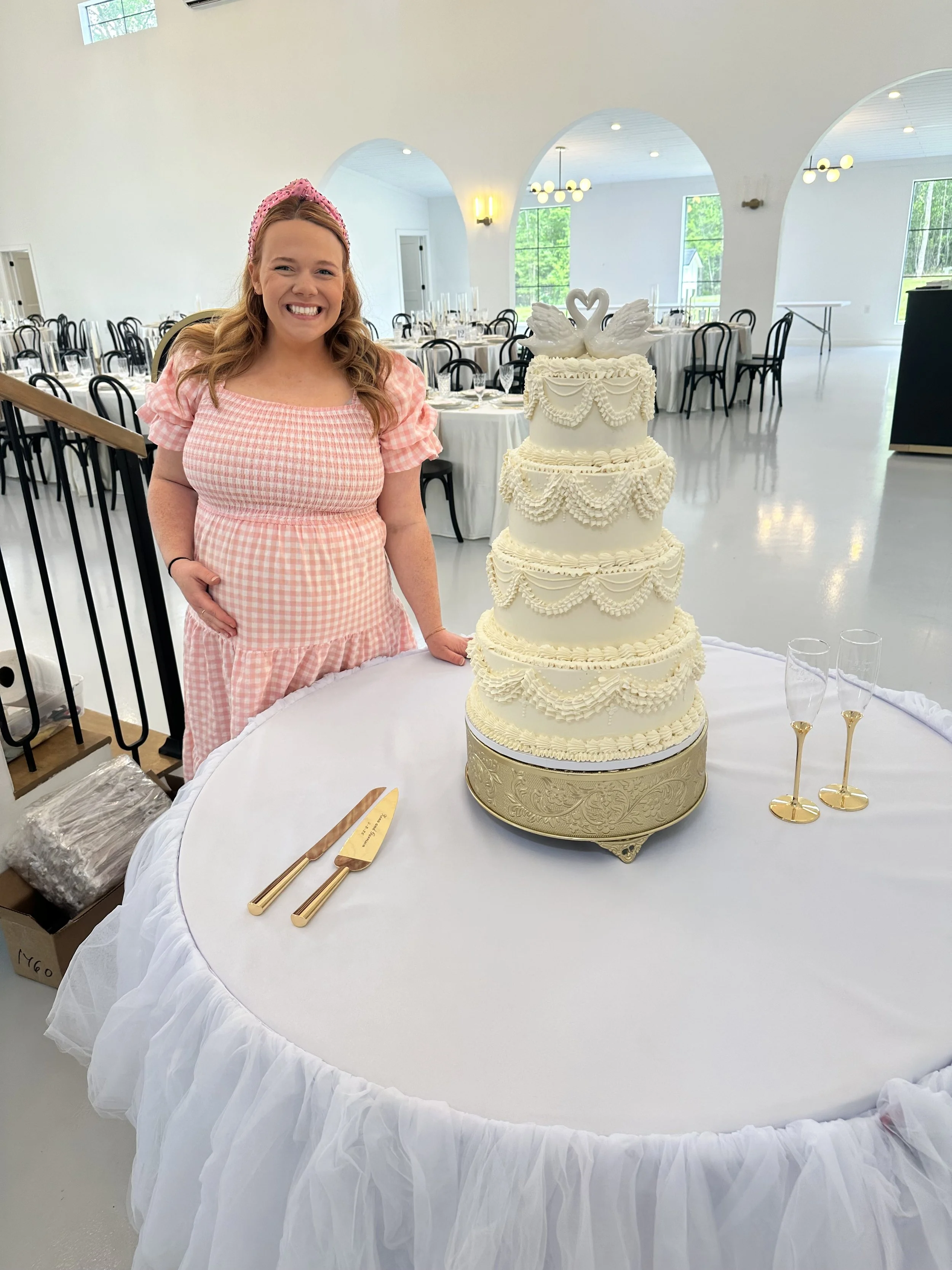 A young woman dressed in a pink gingham dress with a pink headband smiling at a four-tiered white wedding cake decorated with swan toppers and ornate icing piping, on a round table draped with white fabric in a bright, spacious event hall with large 