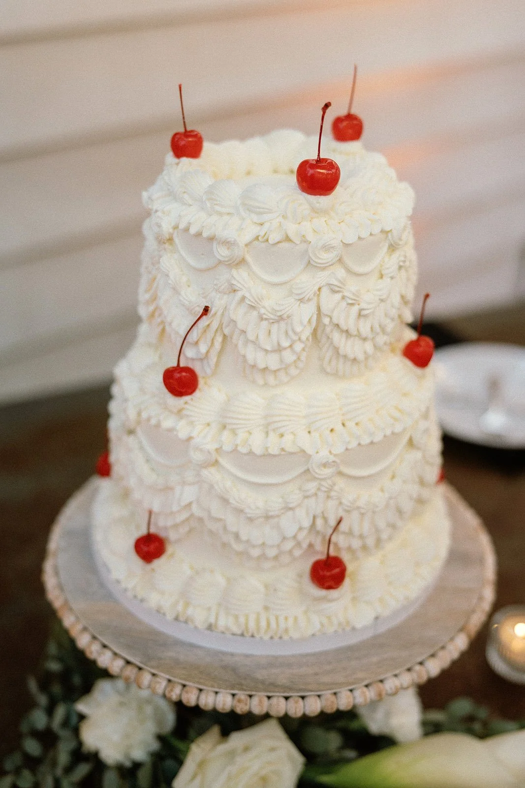 A four-tiered white wedding cake decorated with piped cream and topped with red cherries, placed on a wooden cake stand.