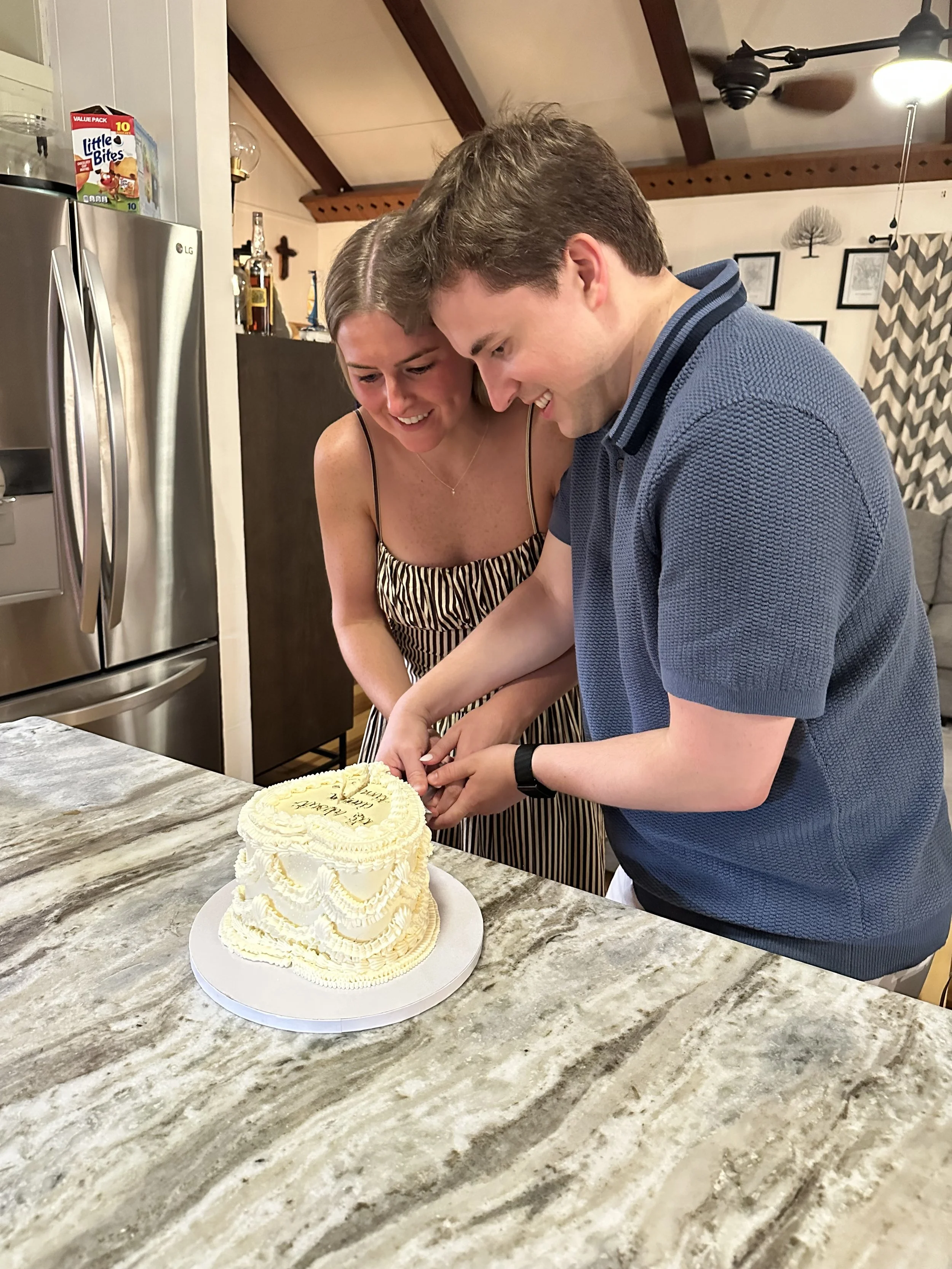A couple is cutting a wedding cake together in a kitchen, smiling and looking at the cake. The cake is a white, multi-tiered cake with yellow accents, placed on a white plate on a marble countertop.