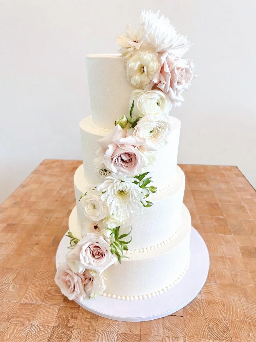 Three-tier white wedding cake decorated with cascading white and blush pink flowers, placed on a wooden table.