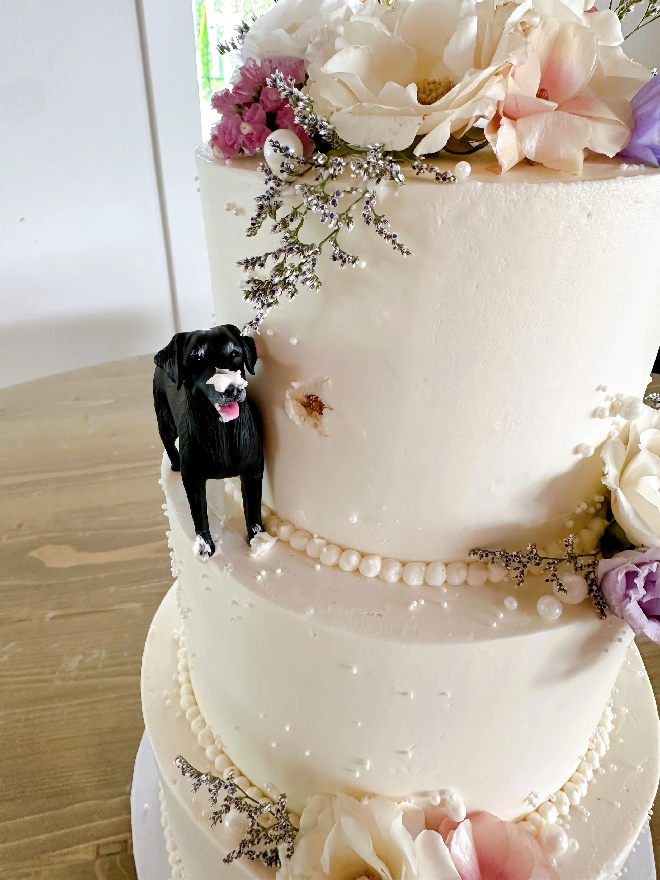 A white, multi-layered cake decorated with flowers, beads, and a small dog figurine with black fur, a white snout, and pink tongue.