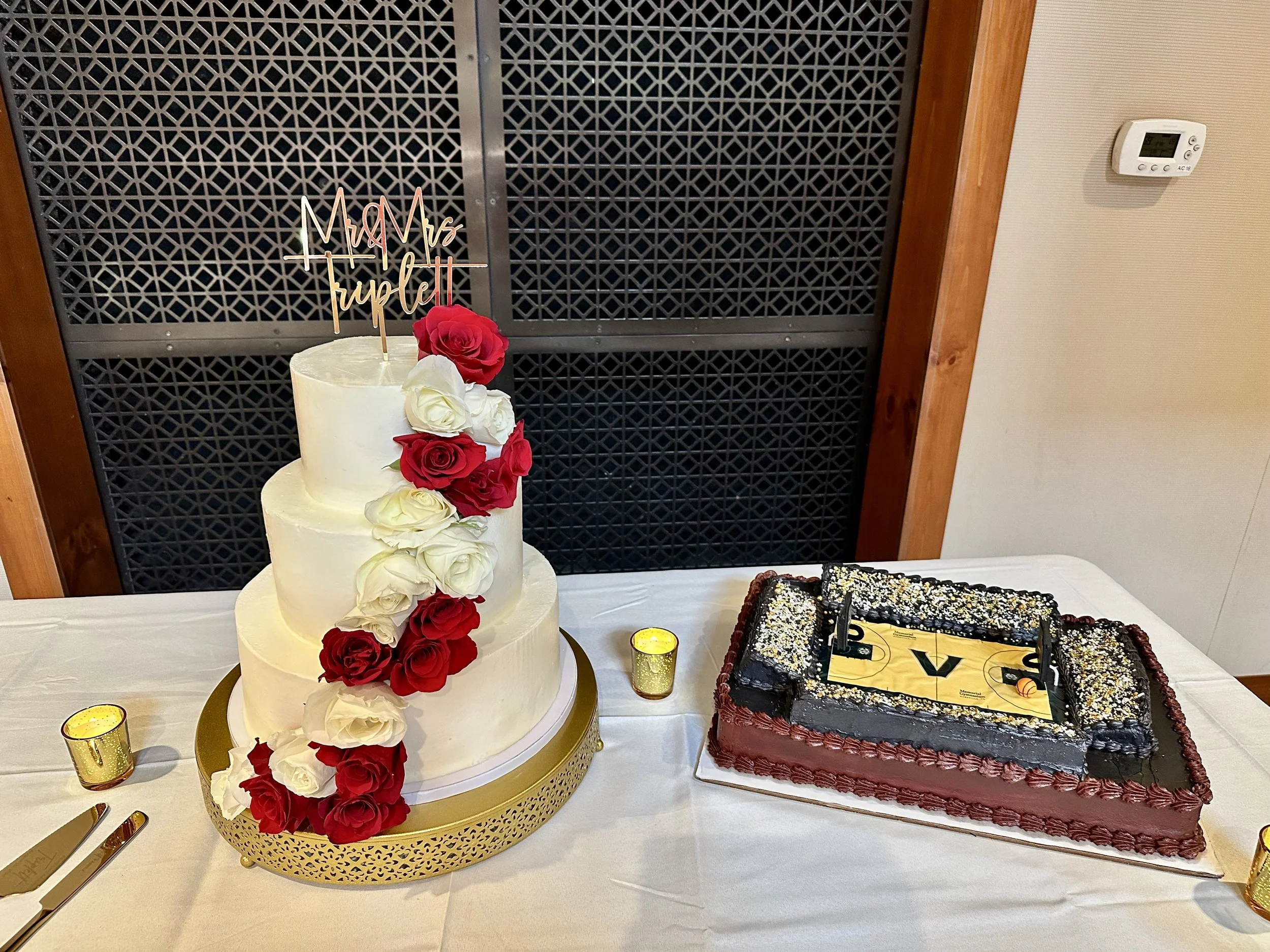 A white, multi-tiered wedding cake decorated with red and white roses and a "Mrs. Trujillo" cake topper, placed on a gold tray. Next to it are two bottles of champagne and a black decorated cake. There are small gold votive candles around the table.