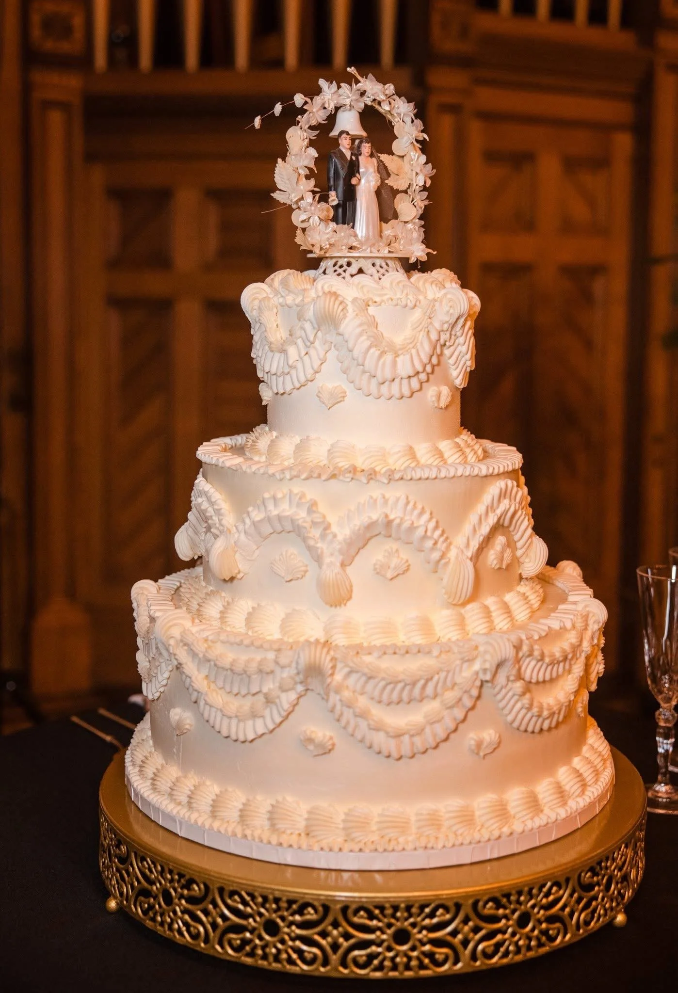 Three-tier wedding cake decorated with white frosting and shells, topped with a figurine of a bride and groom under a floral arch.