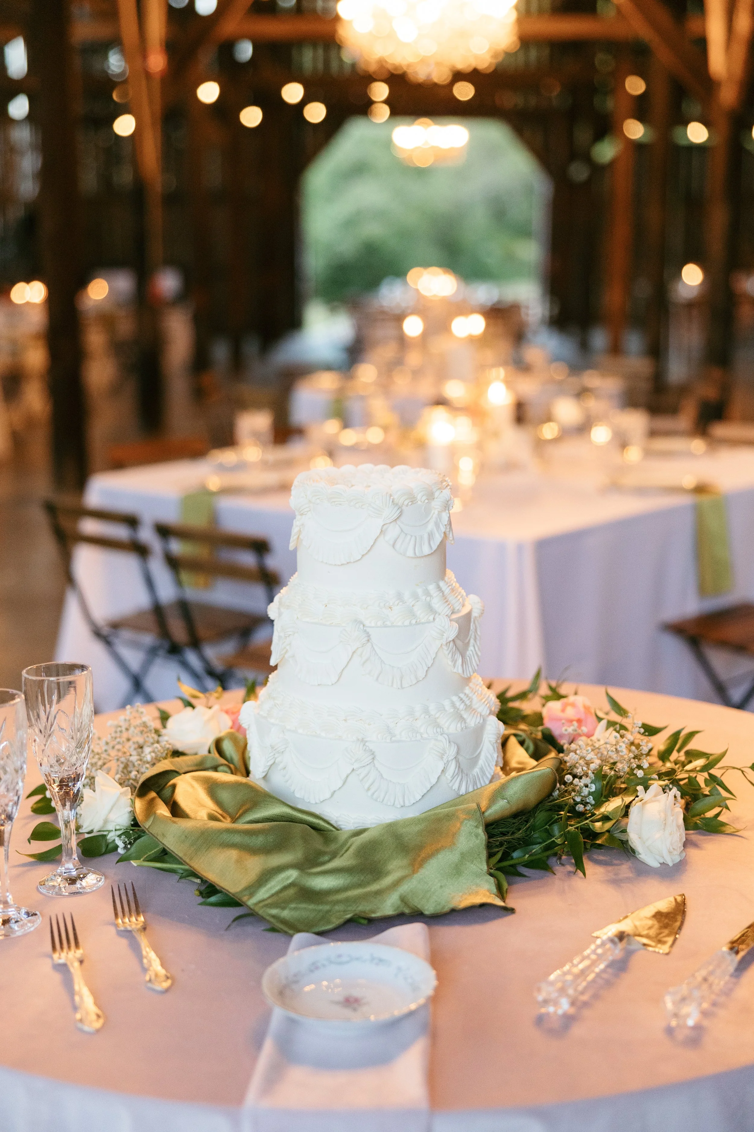 A wedding cake decorated with white icing and ruffled details, surrounded by flowers and greenery, on a table set for celebration in a rustic barn with string lights.