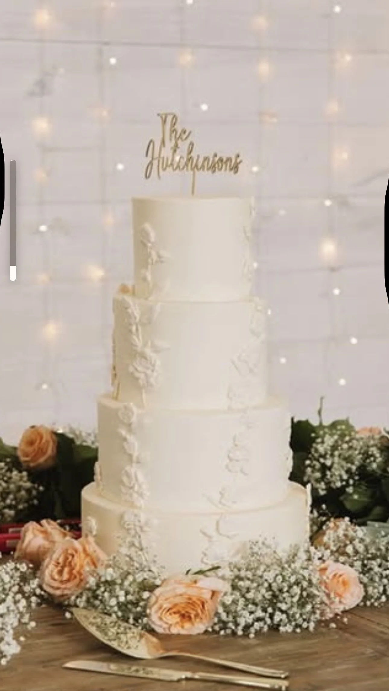 A four-tier white wedding cake decorated with white flowers, topped with gold text that says "The Hutchings." Surrounding the cake are peach-colored roses, white baby's breath, and greenery, with a gold spoon and knife in the foreground.