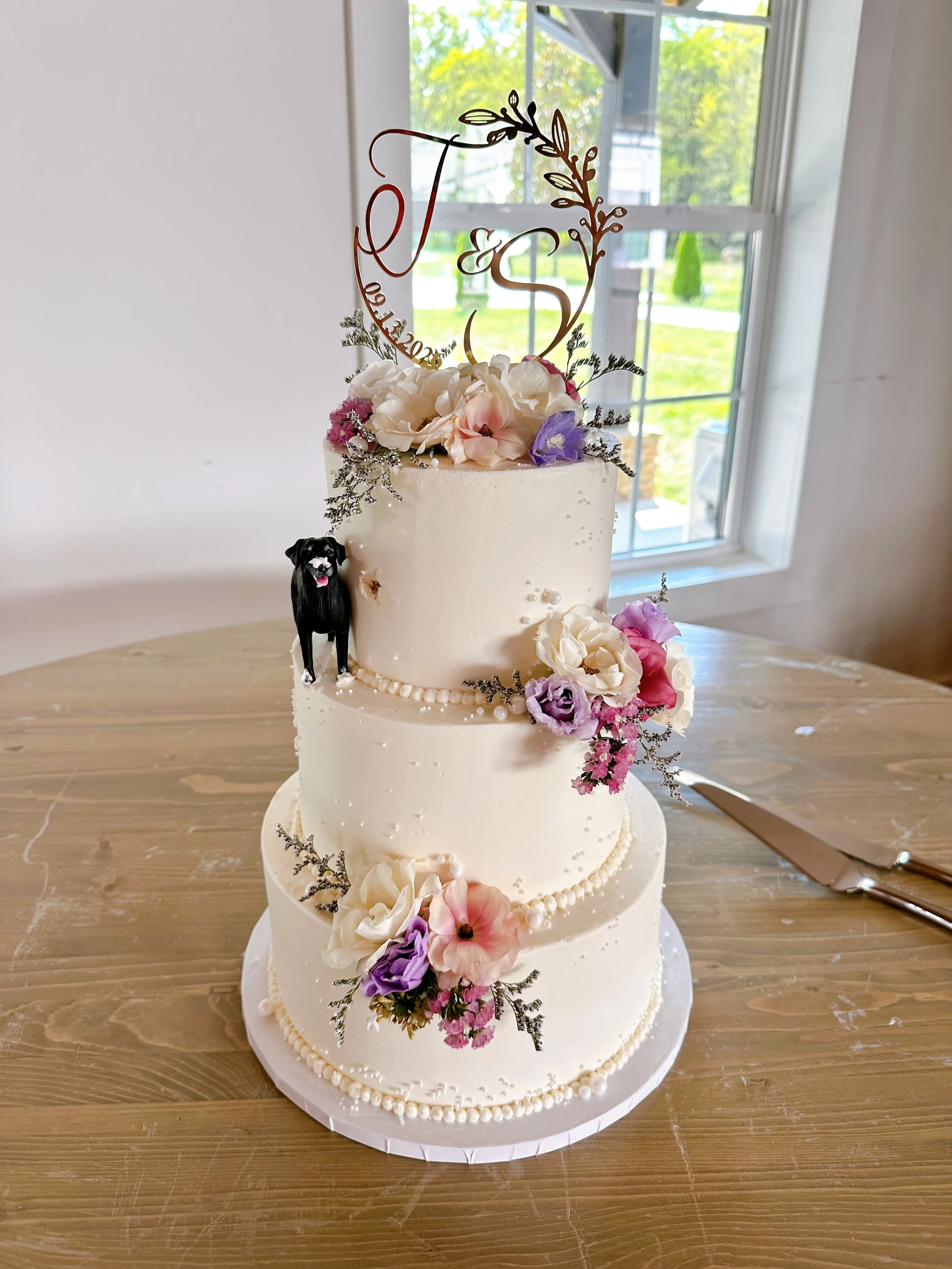 Three-tiered wedding cake decorated with white, purple, and pink flowers, green foliage, and a gold topper featuring the initials T and S with a floral design, on a wooden table near a window.