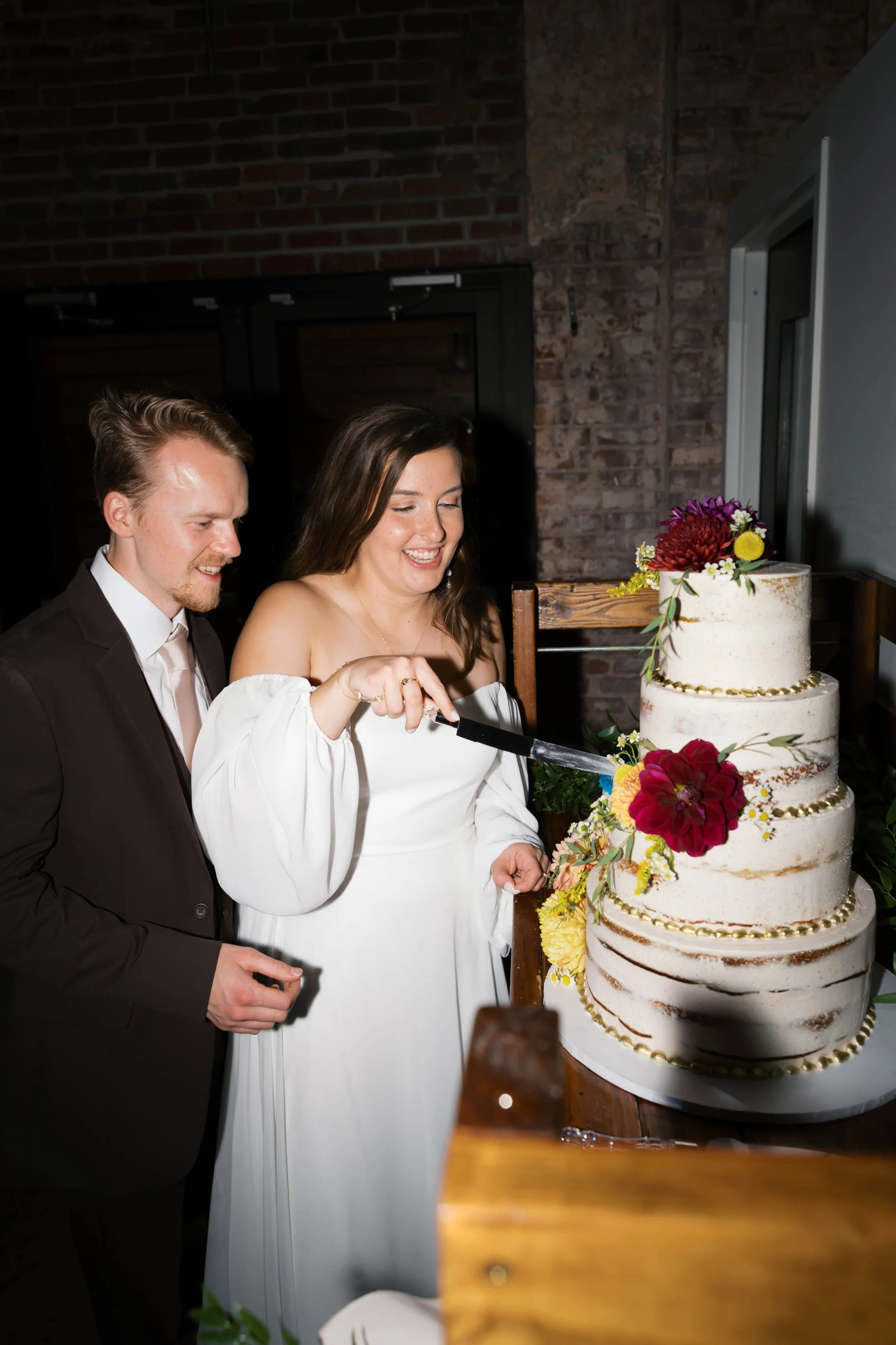 A bride and groom cut a wedding cake together, smiling. The cake is decorated with flowers and gold accents, placed on a table with a rustic brick wall background.