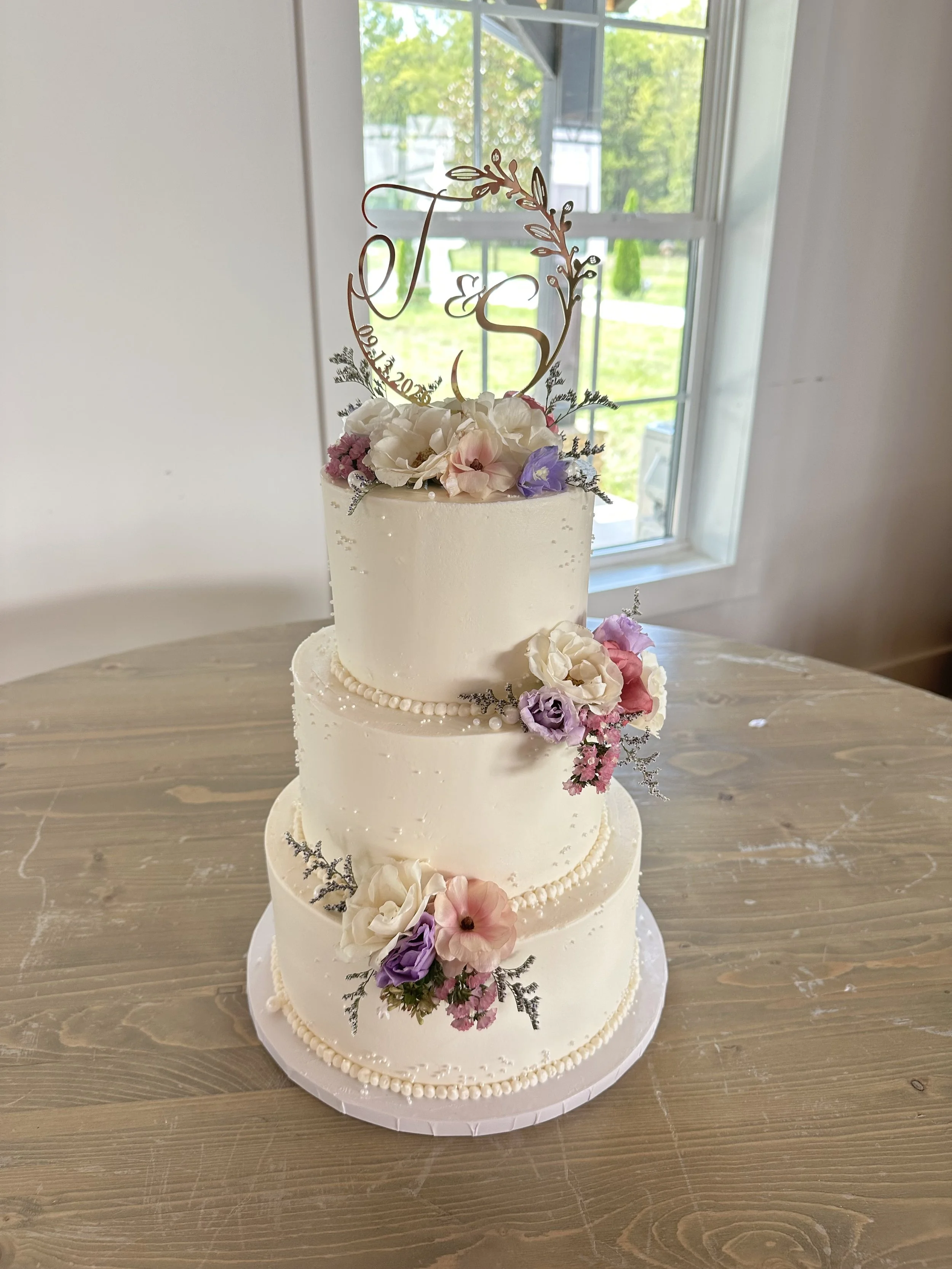 A multi-tiered white wedding cake decorated with pink, purple, and white flowers, topped with a gold monogram cake topper featuring the initials 'T & S' and greenery, sitting on a wooden table near a window.