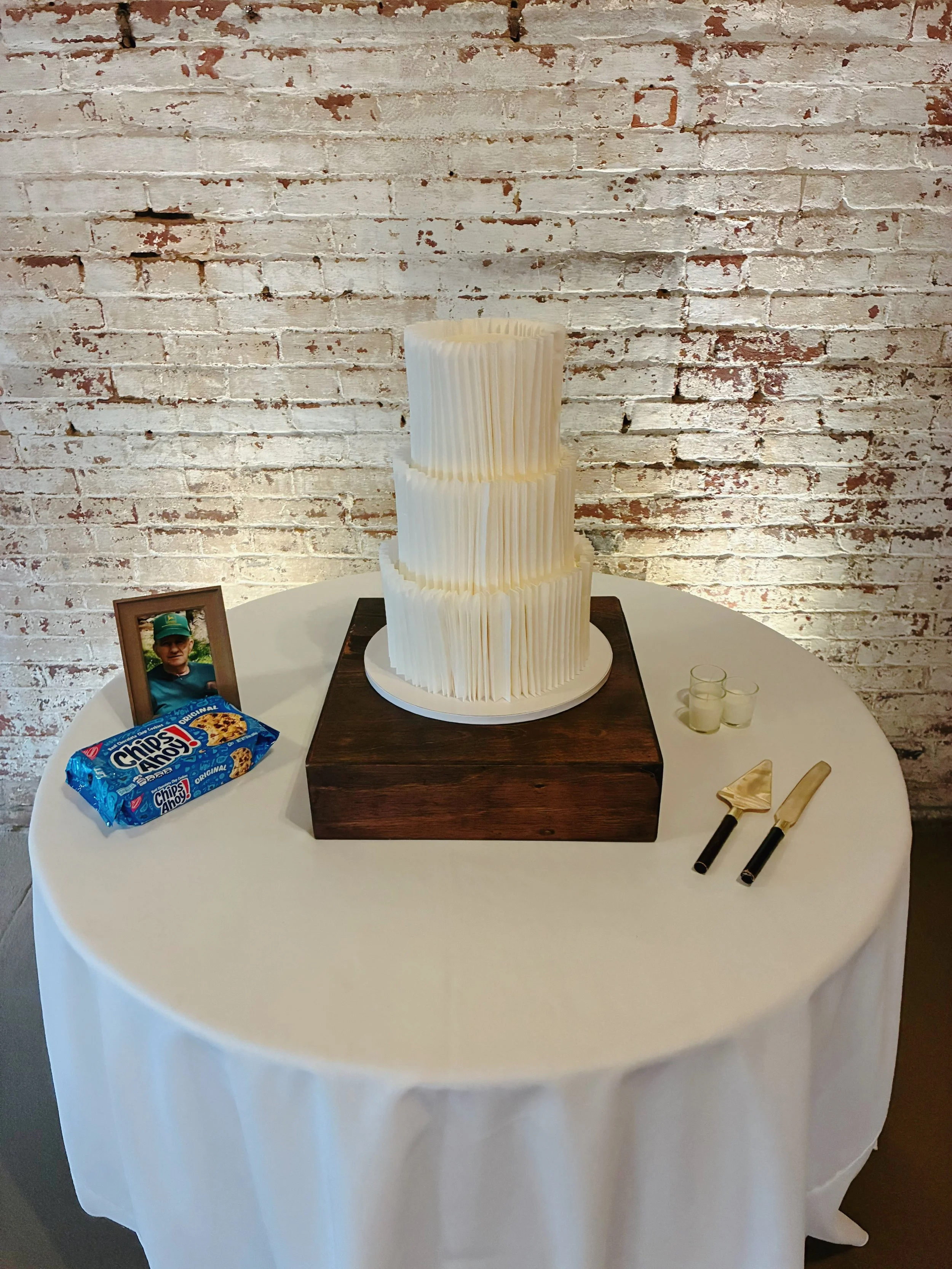A white, tiered cake with vertical ridges sits on a wooden platform on top of a round table covered with a white tablecloth. To the side of the cake, there are a framed photograph of a young man and a pack of Chips Ahoy! cookies. There are three smal