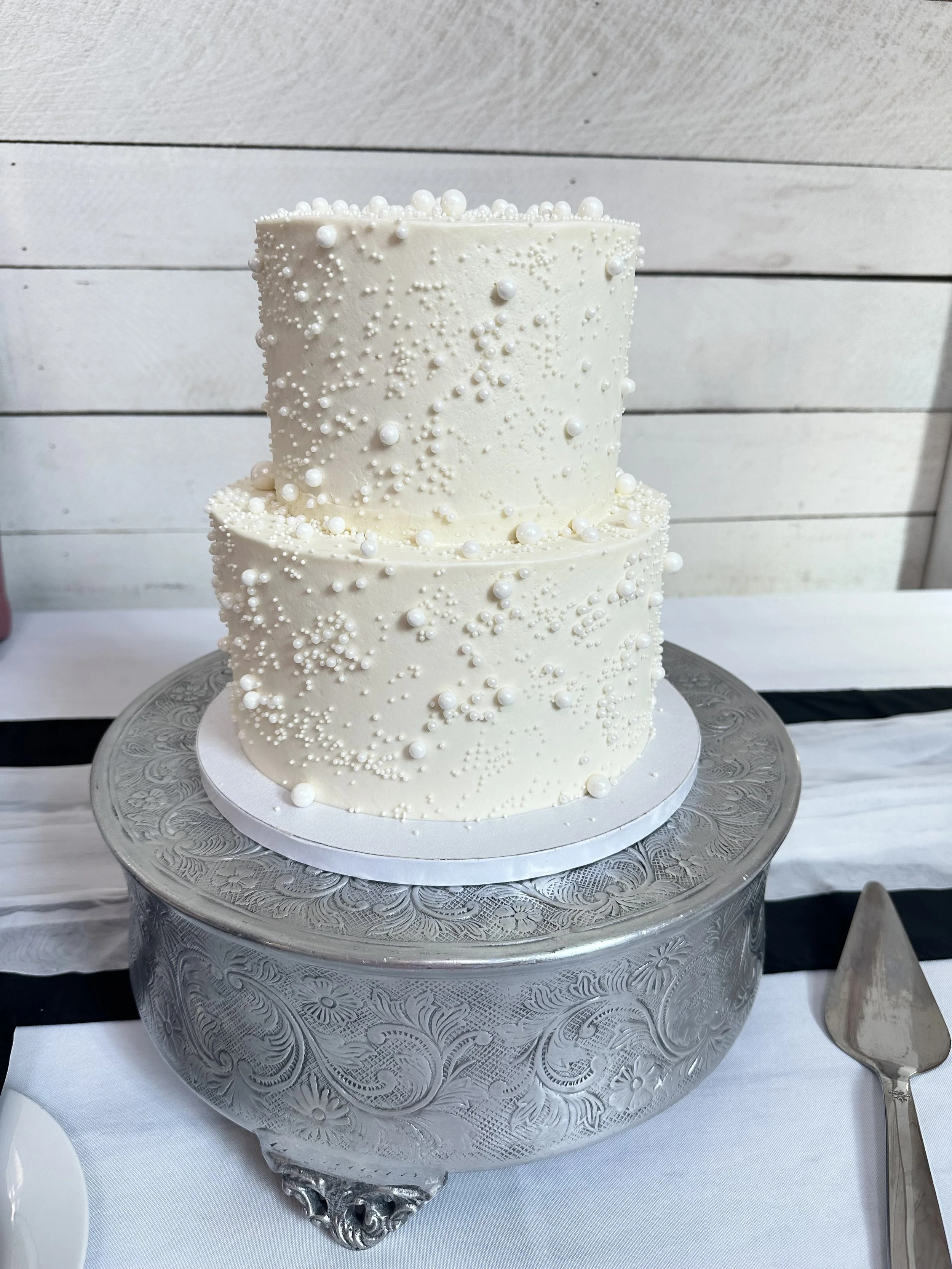 Two-tier white wedding cake decorated with white pearls and small edible beaded decorations, placed on a silver cake stand.