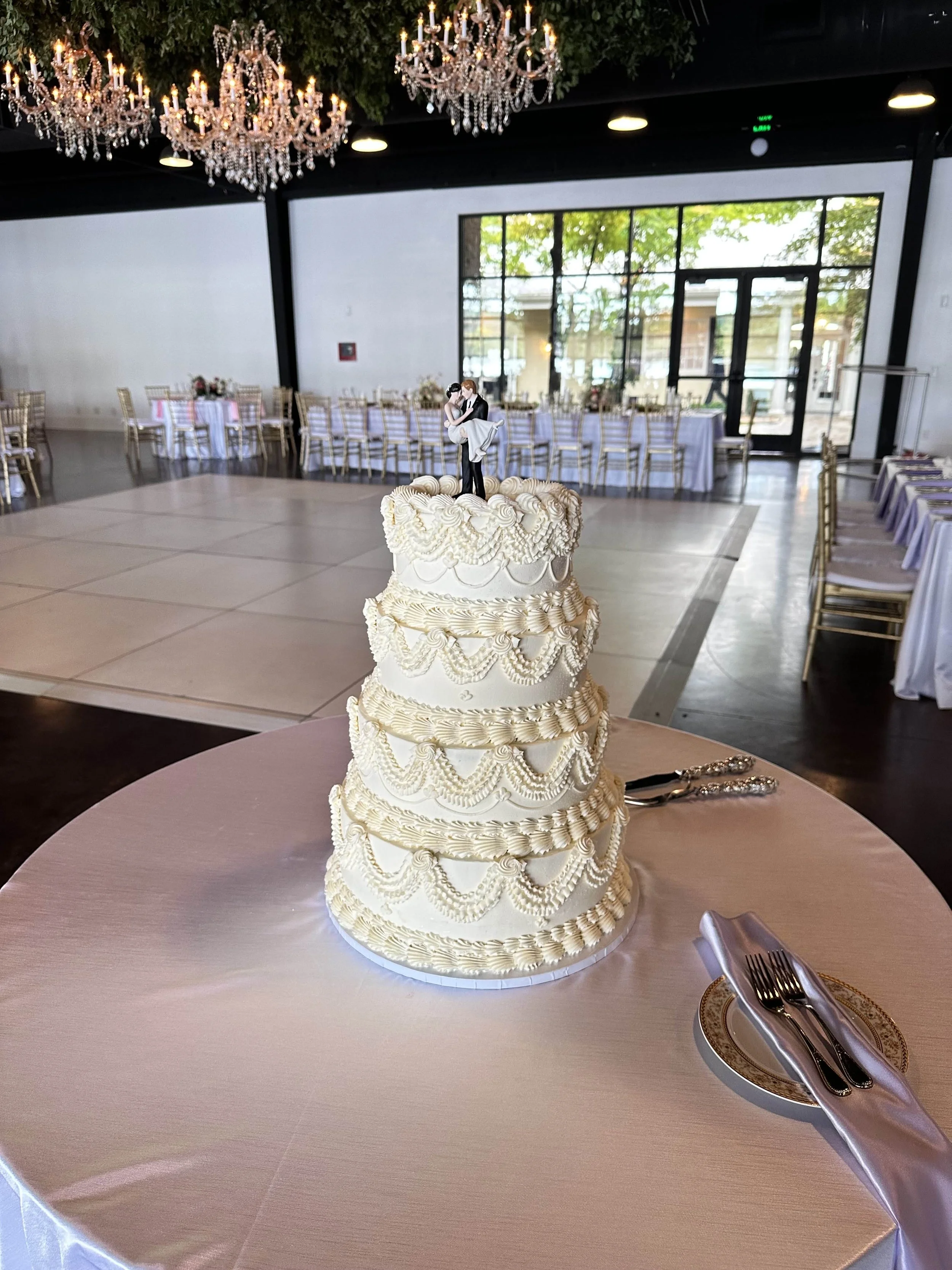 Elegant wedding cake with four tiers decorated with piped icing detailing, topped with a bride and groom cake topper, on a round table with a silver plate and utensils in a decorated reception hall with chandeliers and long dining tables.