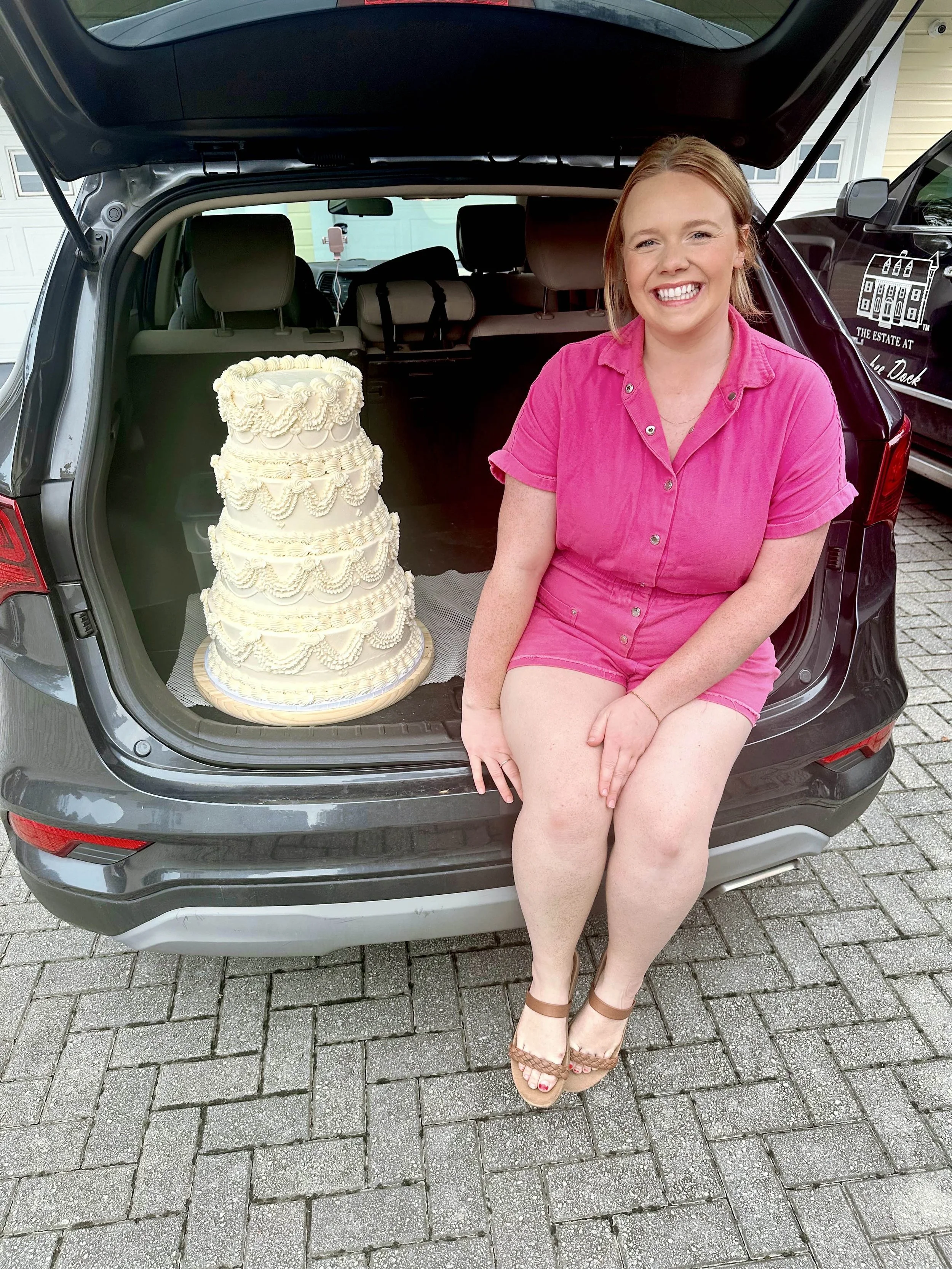 A woman sitting on the edge of an open car trunk next to a tall, multi-tiered white wedding cake, smiling at the camera.