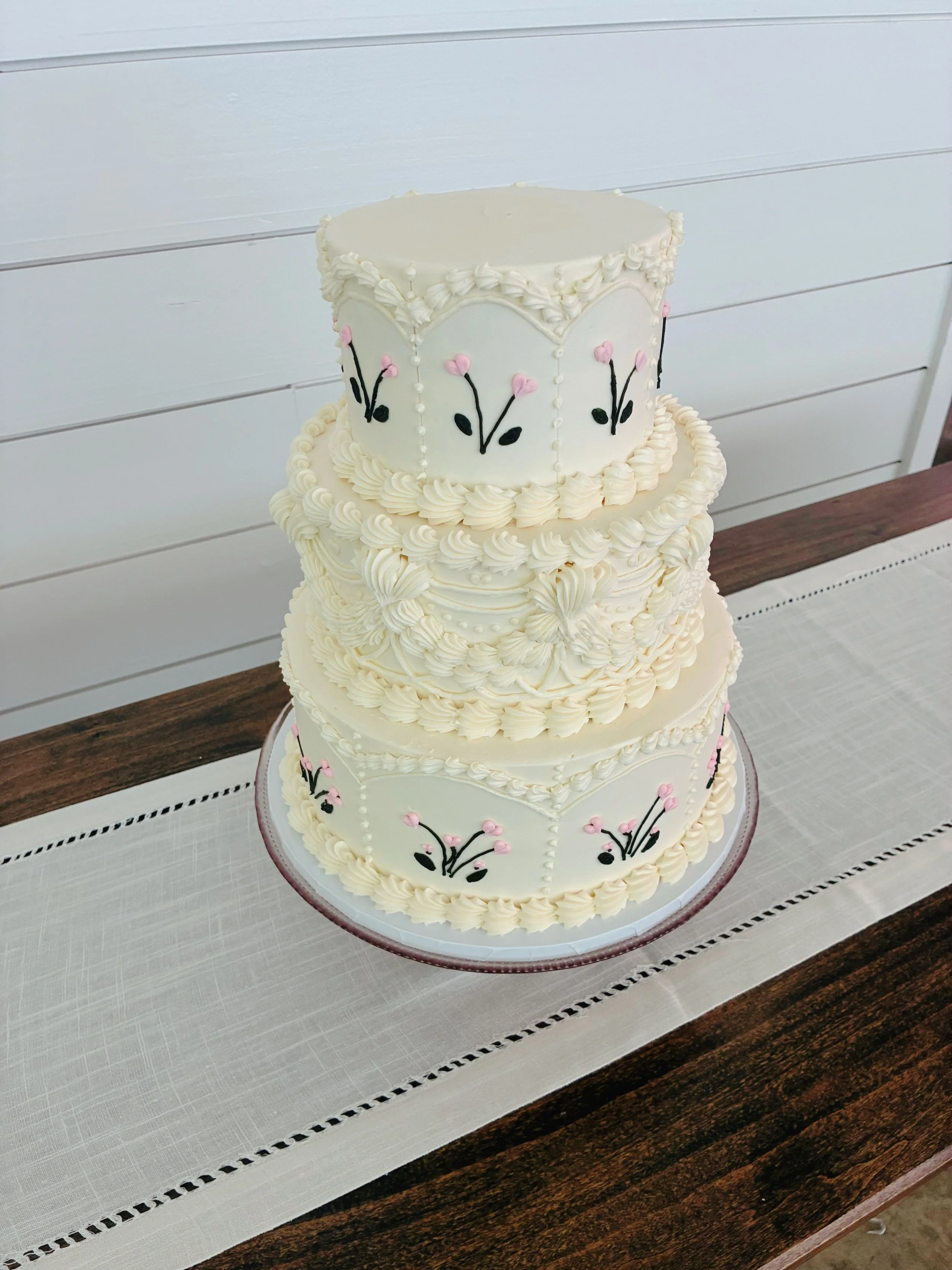 A multi-tiered white wedding cake decorated with pink and black flower designs, on a silver base, placed on a table with a white tablecloth.