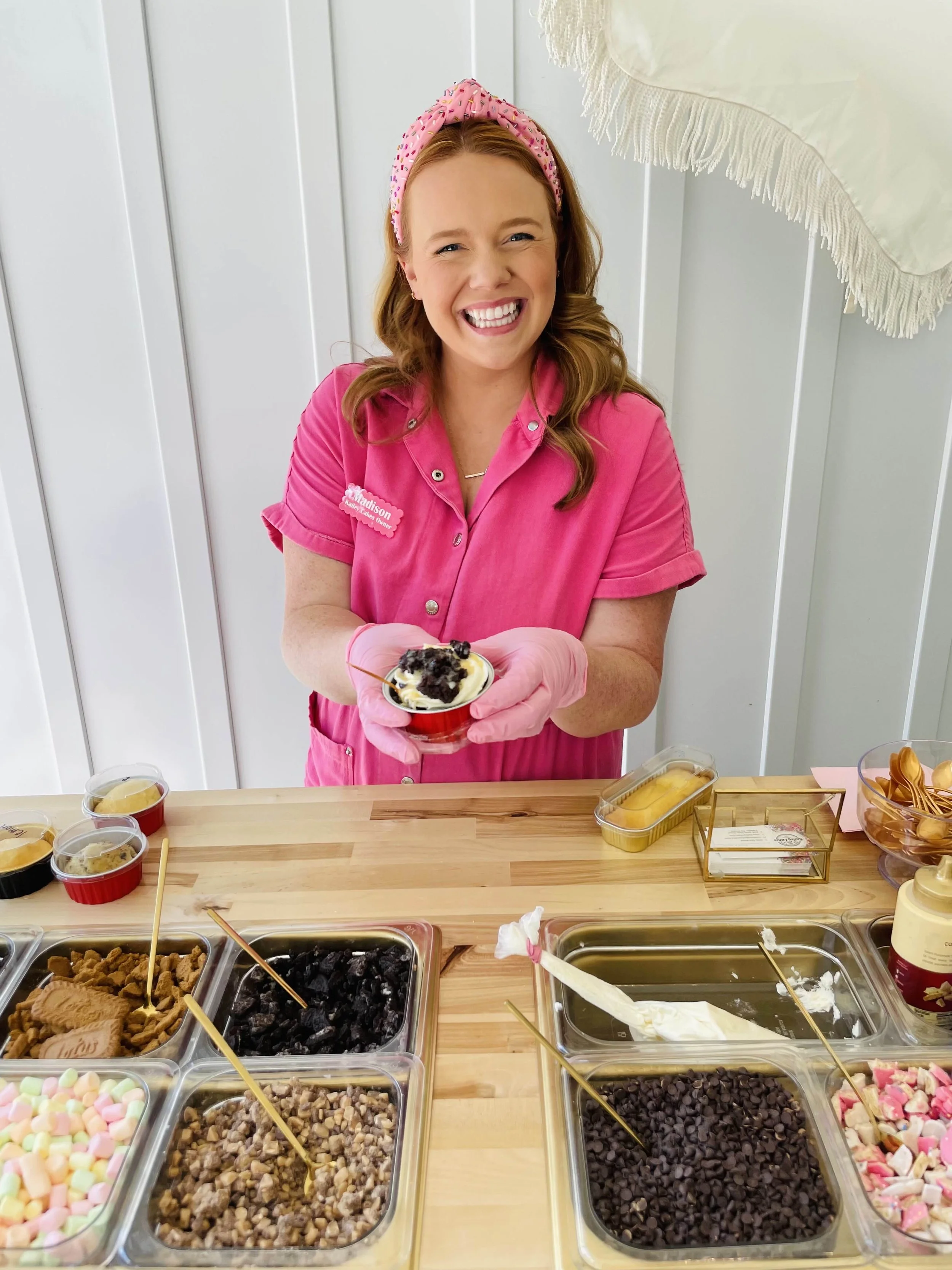 A woman in a pink uniform serving ice cream in a small cup at a dessert station, with various toppings and mix-ins on the table.